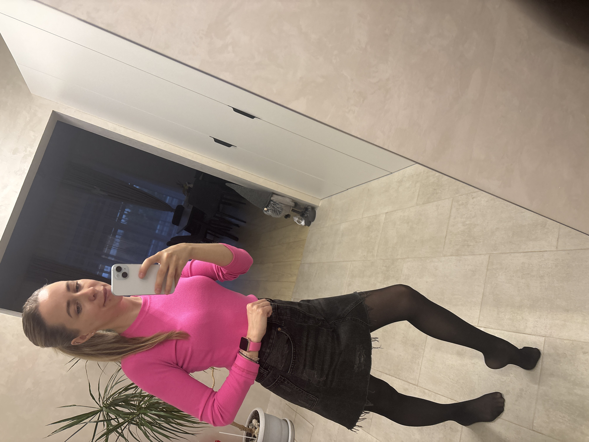 A woman is taking a selfie in what appears to be an indoor setting. She is wearing a bright pink long-sleeved shirt, a black denim skirt, and black tights. She is holding a white smartphone in her right hand, using it to capture the image. The background features a modern-looking interior. There's a white cabinet or wardrobe with sleek handles and a glimpse into an adjacent room with what seems to be a dining area. A potted plant is in the foreground on the left side of the frame. The location cannot be determined from the image, nor is there enough information to confidently place the image in Chaikovsky, Russia.