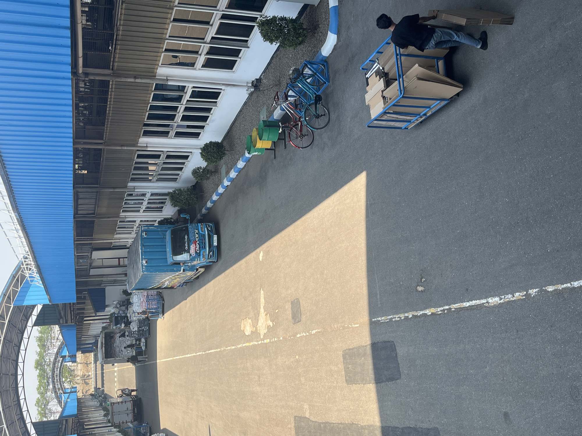 This image depicts an outdoor loading dock or industrial area, possibly within the city of Pelang, Indonesia, characterized by a long, paved pathway bordered by buildings with blue roofs and white facades. Several trucks are present, including a blue truck with the logo "devani" and other delivery vehicles, some loaded with goods. People are visible interacting with the vehicles and cargo, with one individual pushing a blue cart filled with cardboard boxes. The scene is bathed in sunlight, casting shadows across the pavement. Several bicycles are parked near a row of green and yellow trash bins, suggesting a functional workspace. Trees are sparsely planted along the perimeter of the buildings. The overall impression is one of a busy industrial or logistics hub.