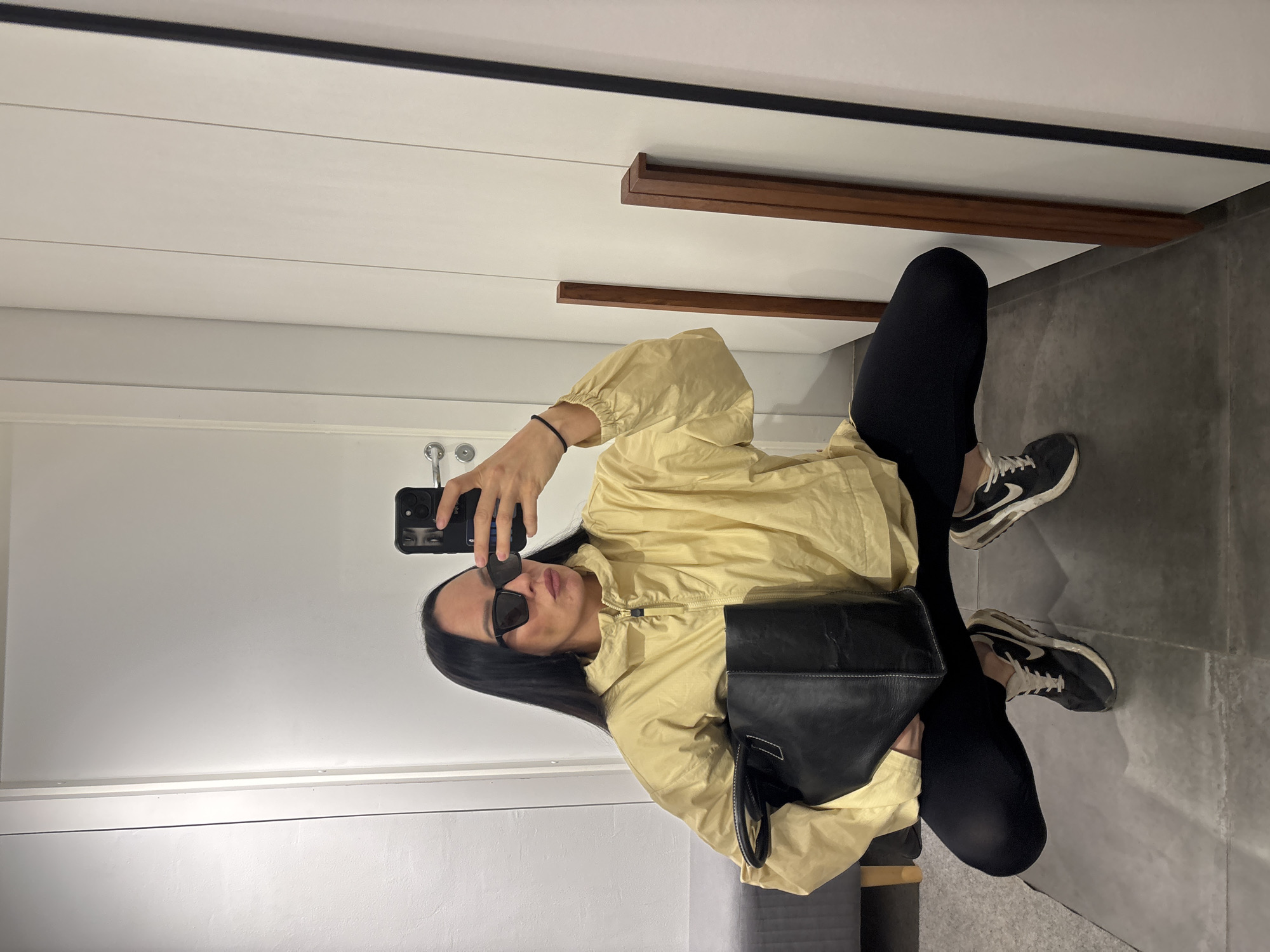 A woman is taking a selfie in a room. She is wearing black leggings, a light yellow jacket, black sneakers, and sunglasses. She is holding a black bag. The background shows a white wall with a black and brown accent and a grey floor. There is no indication that this image was taken in Chaikovsky, Russia.