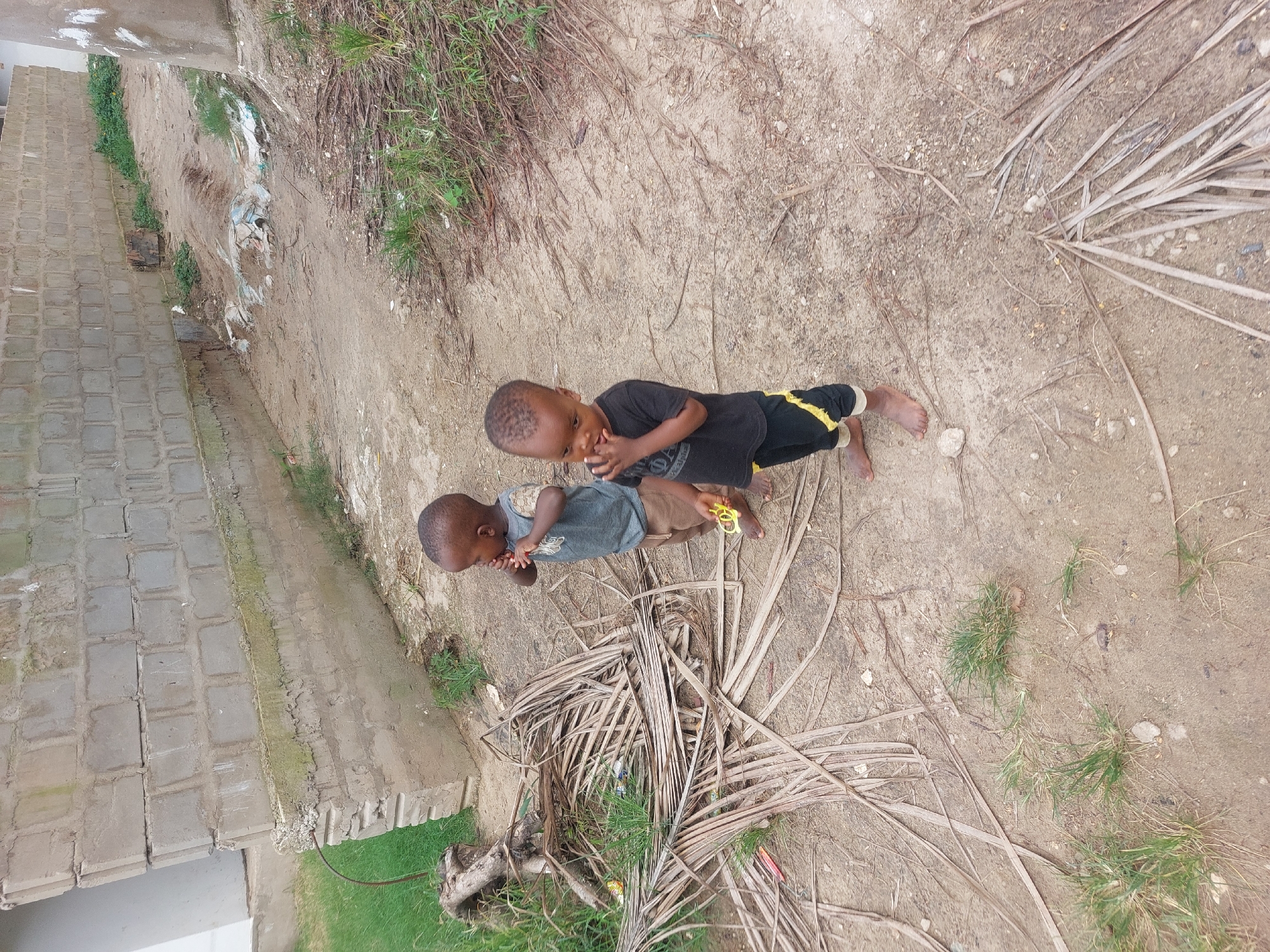 Two barefoot children are standing outdoors on a dirt ground strewn with dry leaves and sparse patches of grass. One child is wearing a black shirt and black pants with yellow stripes, while the other is in a gray shirt and brown pants. They appear to be looking at something in their hands. The background includes a brick building, some green vegetation, and more dry leaves. The location is Dar es-Salaam, Tanzania.