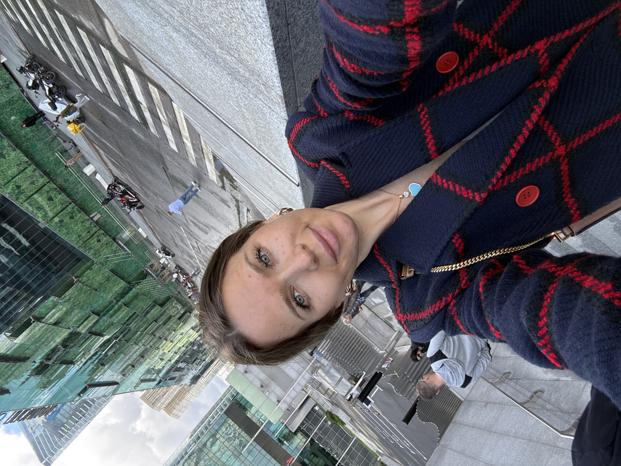 In Moscow, Russia, a woman with light blue eyes and brown hair takes a selfie in front of a street with a pedestrian crossing, several motorcycles, and modern green-tinted glass buildings. She wears a dark blue coat with red plaid accents and a turquoise heart-shaped pendant necklace. In the background, several people are visible on the street, some on foot and others near motorcycles. The sky is cloudy.