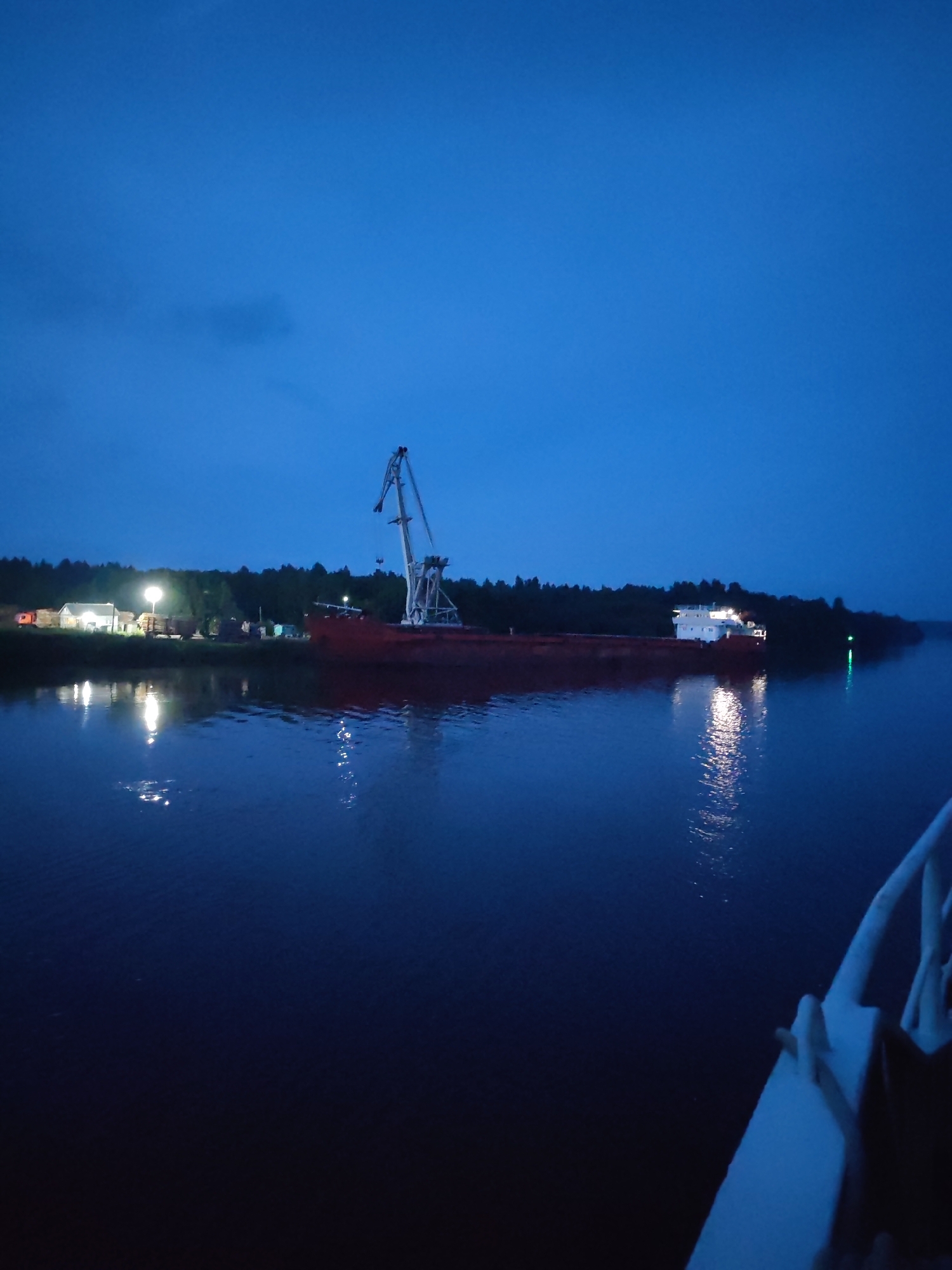 In the twilight hours, a large, red cargo ship is docked on the dark, calm waters of the Svir River at Annensky Most, Russia. A towering, white gantry crane stands at the ready on the shore, silhouetted against the deep blue sky. The riverbanks are lined with a dense forest of trees, their dark forms merging with the approaching night. On the left bank, a cluster of buildings is illuminated by bright lights, suggesting a port or industrial area, with stacks of what appear to be lumber piled nearby. The ship's reflection shimmers on the water's surface, along with the scattered lights from the shore. In the foreground, the white railing of another vessel is visible, hinting at the perspective from which the photograph was taken. The overall scene is one of quiet industry and the transition from day to night.