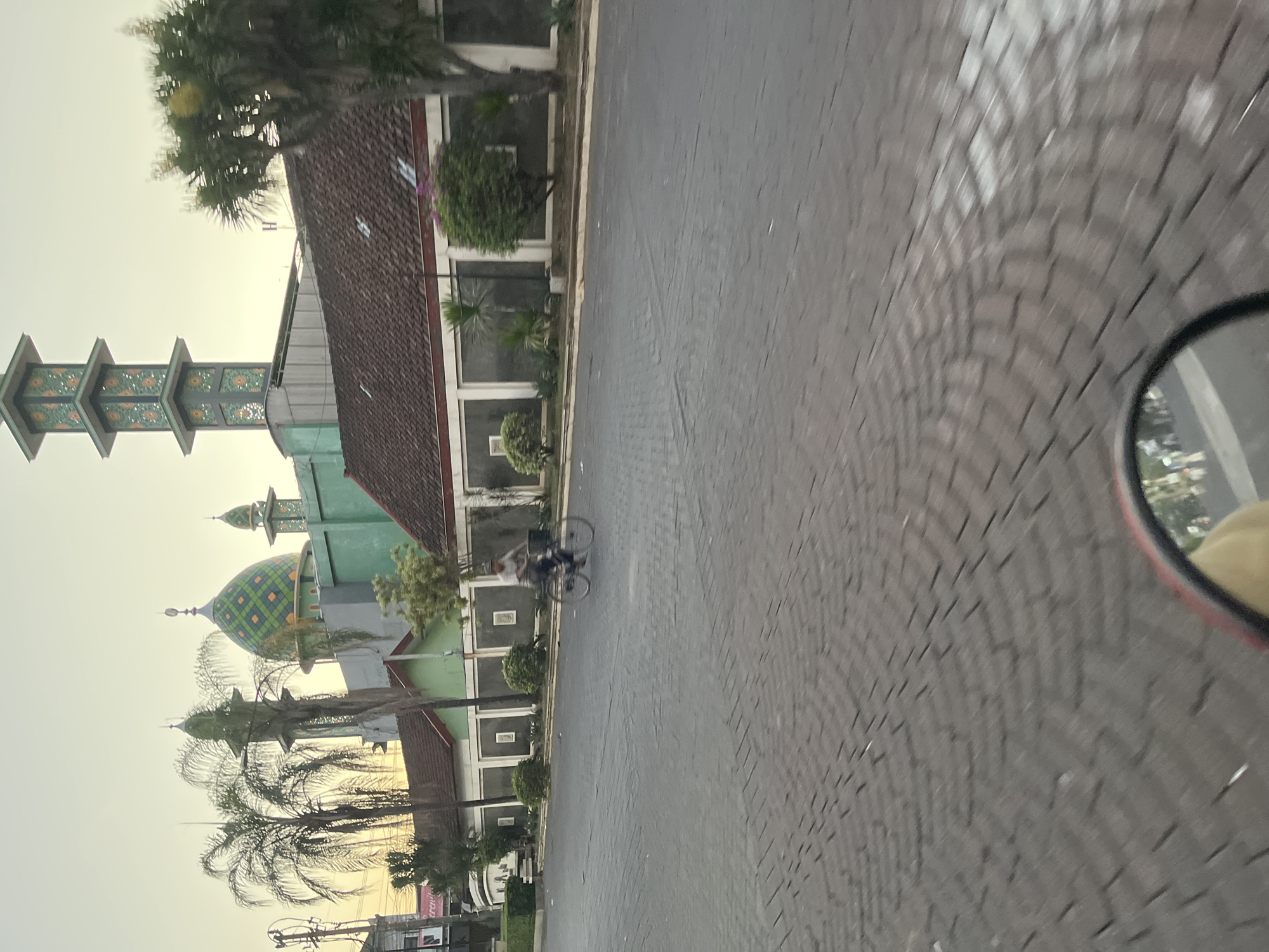 A person rides a bicycle along a paved street in Boro, Indonesia, near a mosque. The mosque has a green dome and minarets adorned with intricate patterns. Palm trees and other foliage surround the mosque and a low wall. The sky is a pale yellow, suggesting either dawn or dusk. The street is constructed of dark gray interlocking pavers. A red-rimmed mirror, likely from a vehicle, is visible in the foreground, reflecting a portion of the scene. The overall impression is a quiet, everyday moment in a tropical urban setting.