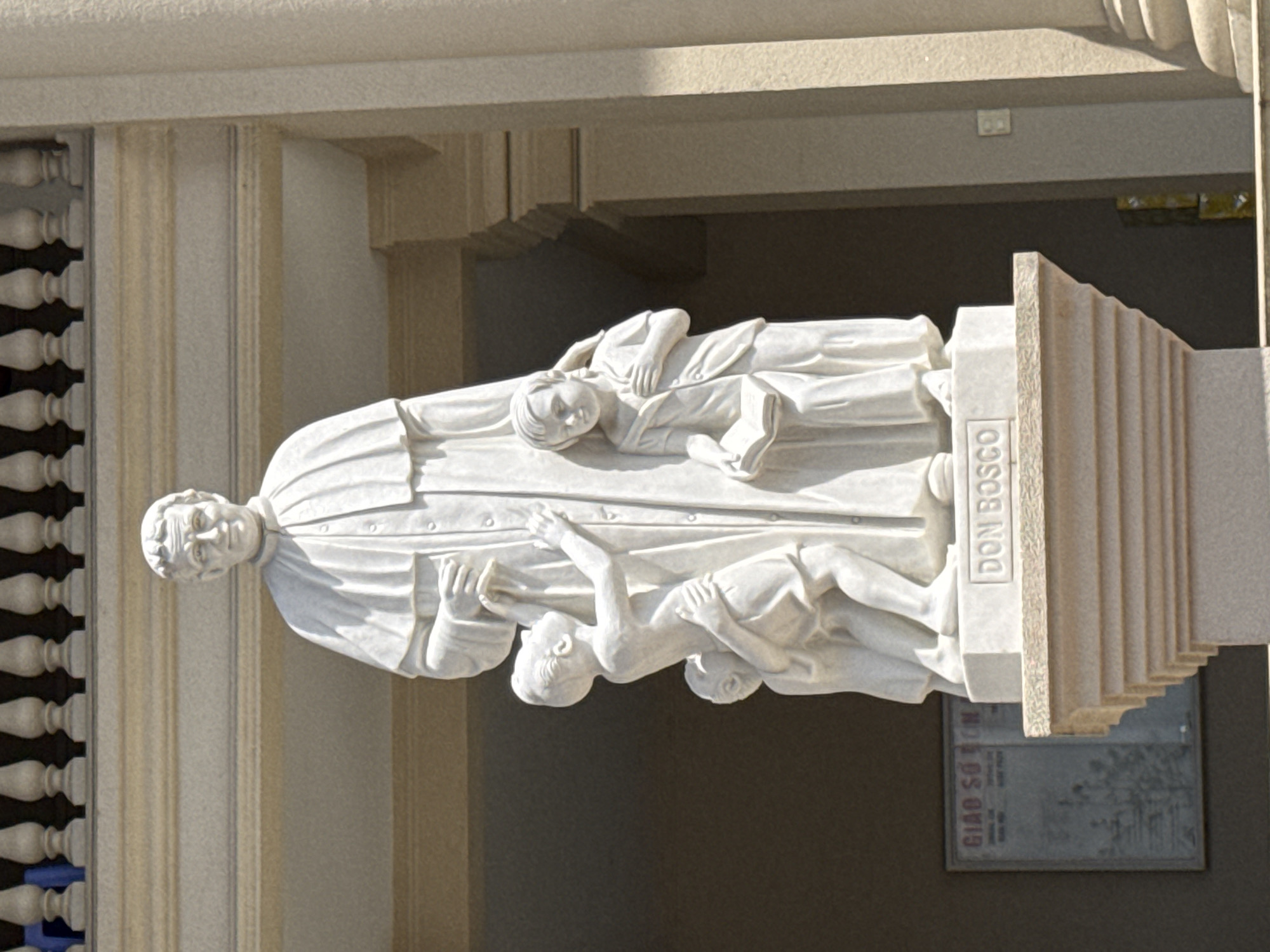 A white marble statue of Don Bosco is situated on the side of a building. The statue depicts Don Bosco with two children clinging to his robes. A plaque bearing the name "Don Bosco" is affixed to the base. The building has stone columns on one side and is located in Da Lat, Vietnam.