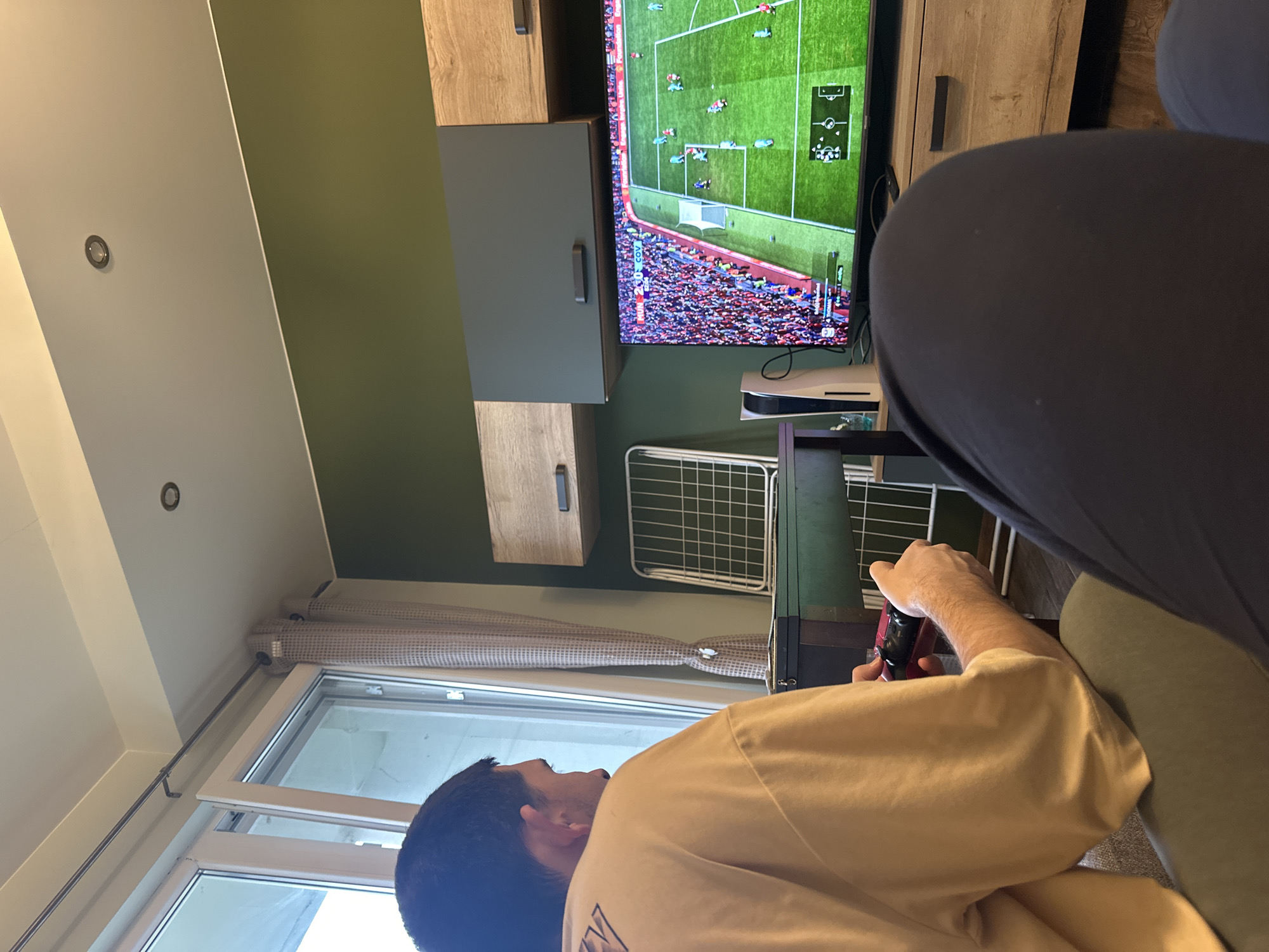 An interior scene in Sofia, Bulgaria, depicts a male individual playing a football video game. The man is seated on a light gray upholstered sofa, viewed from behind and the right side, wearing a loose-fitting light yellow/beige short-sleeved shirt. He holds a red and black game controller with both hands. In the immediate foreground, the lower legs of another person, wearing dark trousers, are visible, indicating a shared viewing or playing experience.
A large flat-screen television dominates the center-right of the frame, displaying a football match with the score "MUN 2 - 0 COV" and an in-game stadium crowded with spectators. A white PlayStation 5 console stands vertically to the left of the television. The television rests on a light wood-grain entertainment console. Above the television, a wall-mounted unit features two dark gray cabinets with horizontal handles flanked by two matching light wood-grain cabinets.
To the left, a large window, with one pane slightly ajar, allows natural light into the room. A light-colored, patterned curtain is partially drawn. Behind the player, a dark green piece of furniture is visible, with a white, grid-patterned metal drying rack positioned further back against the wall. The wall behind the TV and furniture is painted a dark olive green, transitioning to a light beige/off-white on the upper wall and ceiling. Two recessed spotlights are visible in the ceiling. The floor appears to be light-colored wood.