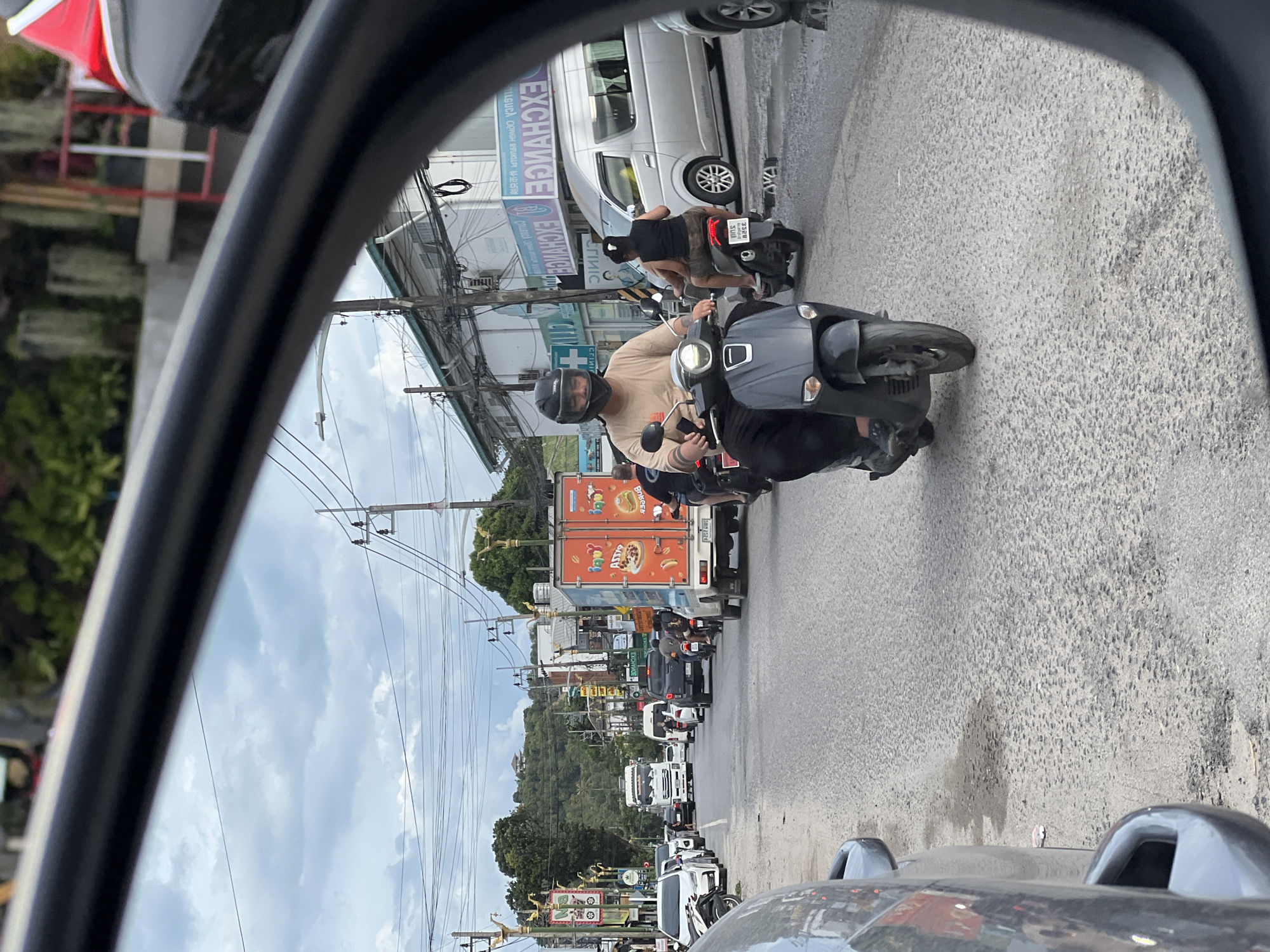 Observed through a car's side mirror, the scene depicts a street in Baan Bang Rak, Thailand. A motorcyclist with a passenger is driving on the right side of the road. Further down the street, there is a line of mixed traffic, including motorcycles, cars, and a delivery truck bearing an advertisement for hamburgers. Buildings are visible along the sides of the street, and utility poles are strung with wires. The sky is overcast.