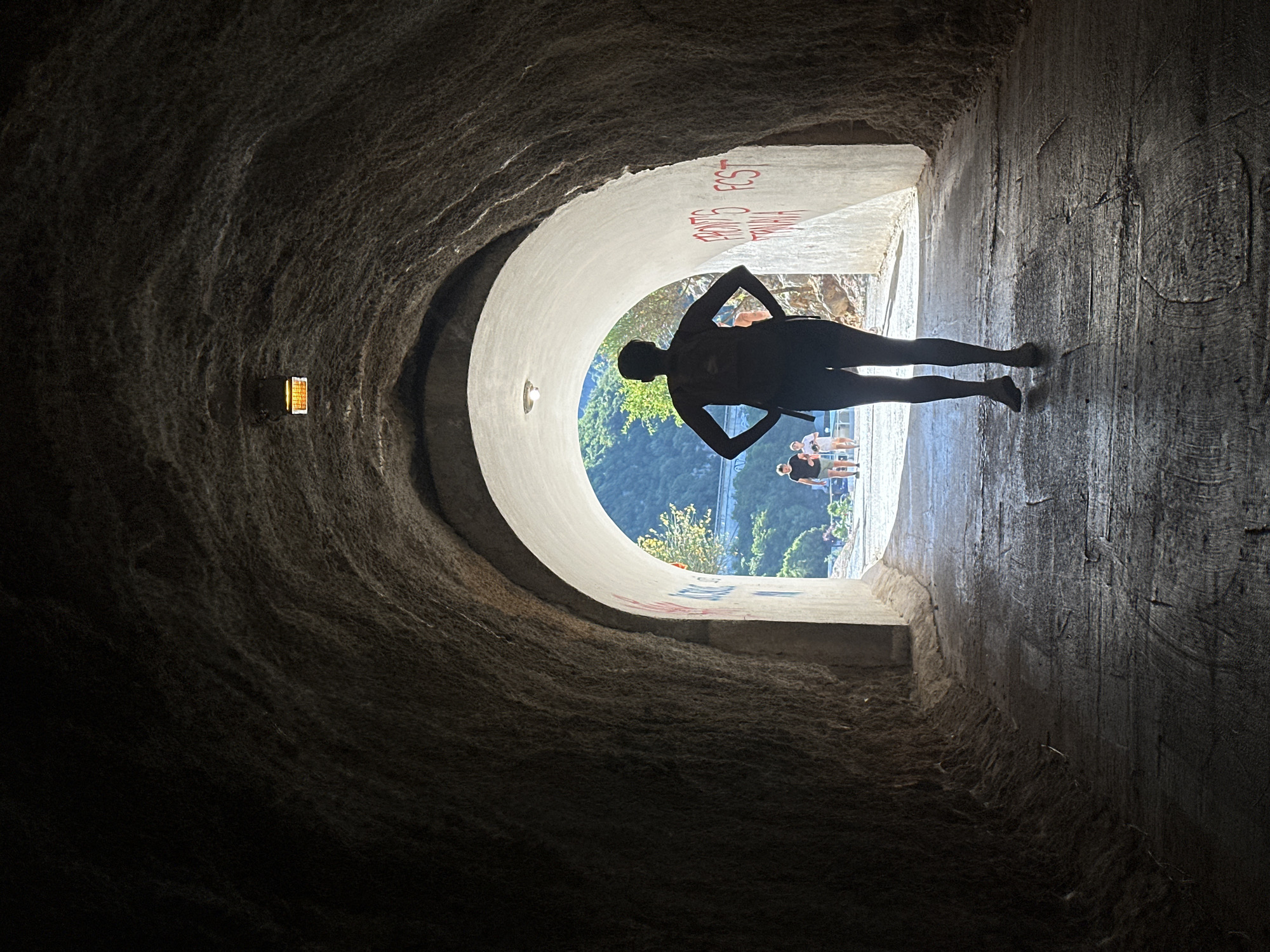 Captured from within a dark, cavernous tunnel, the image shows a silhouette of a person standing at the tunnel's light-filled exit. The tunnel walls are rough and textured, with a small light fixture visible on the left. Graffiti adorns the tunnel walls near the exit. The person's silhouette is positioned in the center of the frame, arms akimbo. Outside the tunnel, a group of people can be seen in the distance against a backdrop of greenery. The scene is located in Bečići, Montenegro.