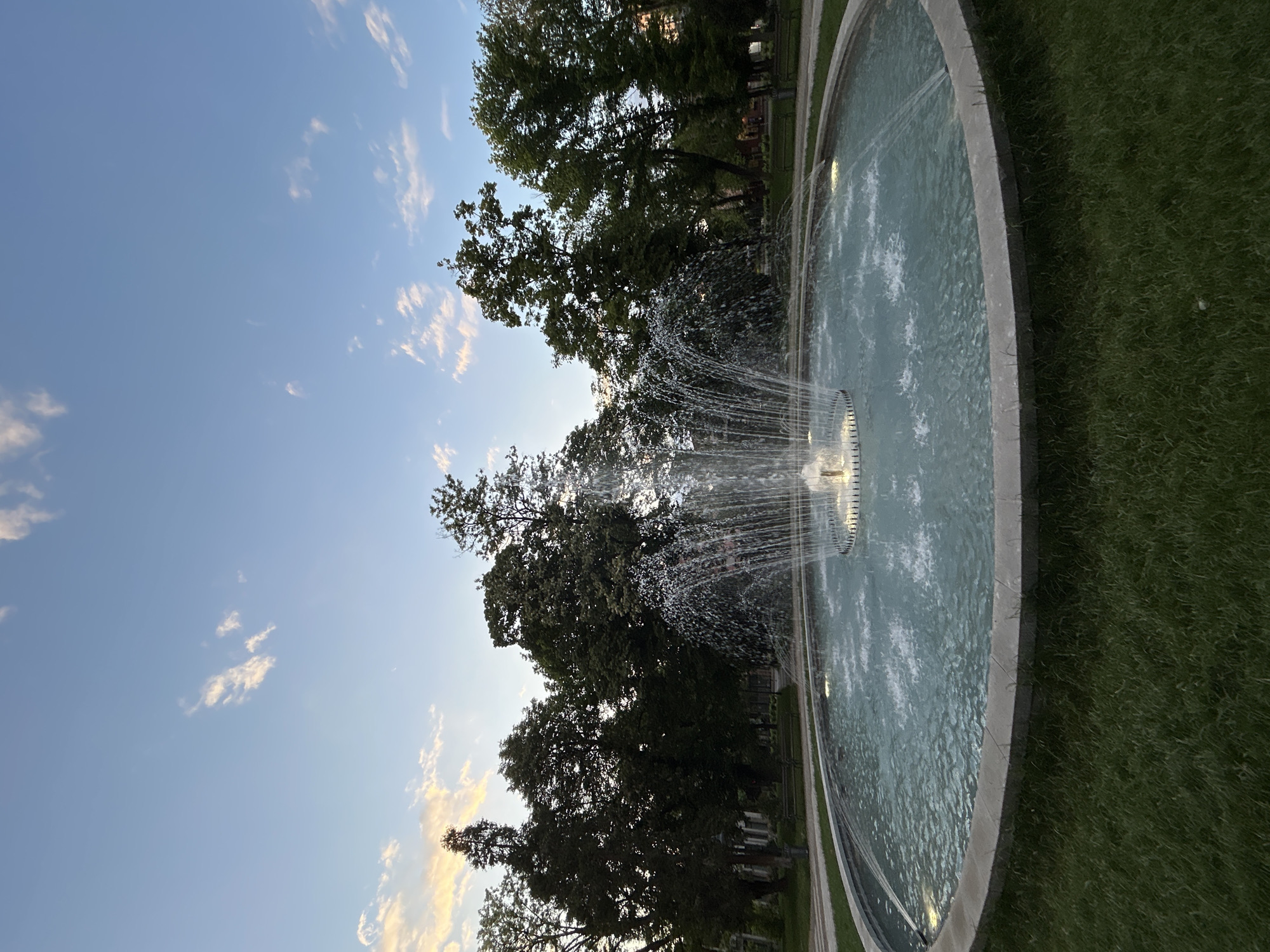 The image shows a circular fountain in a park in Plovdiv, Bulgaria. The fountain is made of concrete and has water spraying from a central structure. The water is clear and reflective. Trees surround the fountain, and the sky is blue with some clouds. Grass is visible on the ground next to the fountain. There are no people visible in the image.