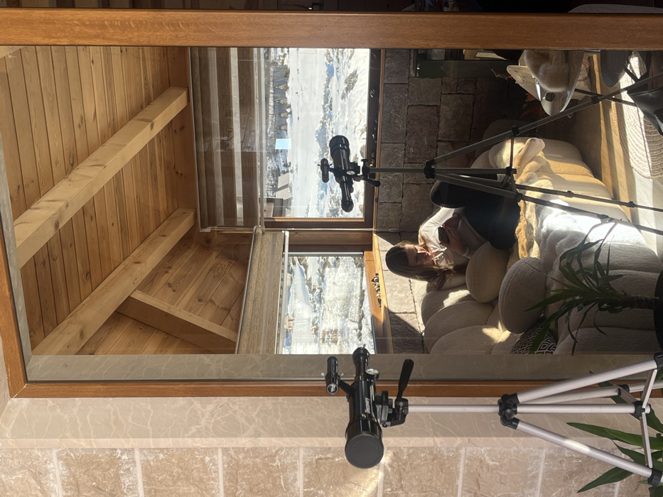The image depicts an interior scene, likely a living space or lounge, situated in Virak, Montenegro, characterized by a panoramic view of a snow-covered landscape. The perspective is through an architectural opening, framed on the left by a light-colored, marble-patterned tiled wall and on the top and right by wooden beams and frames.
In the immediate foreground, to the left, a black telescope mounted on a silver tripod is partially visible. Inside the room, a young adult female with light brown hair is seated on a plush, light-beige sofa. She is wearing a white long-sleeved top and dark trousers, and is actively engaged with a mobile phone held in both hands. Her head is bowed, directing her gaze towards the phone. The sofa is adorned with several cushions, including a round beige one and a black-and-white patterned one, and is partially covered by a white, textured throw or rug. Sunlight streams into the room from the right, casting distinct shadows on the sofa and the light-colored floor. A small potted plant with green leaves is positioned on the floor in front of the sofa.
Further into the room, another identical black telescope on a silver tripod stands to the right of the seated female, positioned near a large window. To the far right, a small round table holds a light-colored, fluffy object.
The room's interior features include a natural wood ceiling with exposed beams. The lower walls are constructed from rough-hewn, light-gray stone blocks. Large windows provide an expansive view of the exterior, which is entirely covered in bright white snow. Beyond the snow-covered ground, several small, triangular-roofed structures, also dusted with snow, are visible in the distance. The windows are fitted with horizontally striped, zebra-style roller blinds, partially lowered. The outdoor scene suggests a cold, clear, and sunny day, contrasting with the warm, illuminated interior.