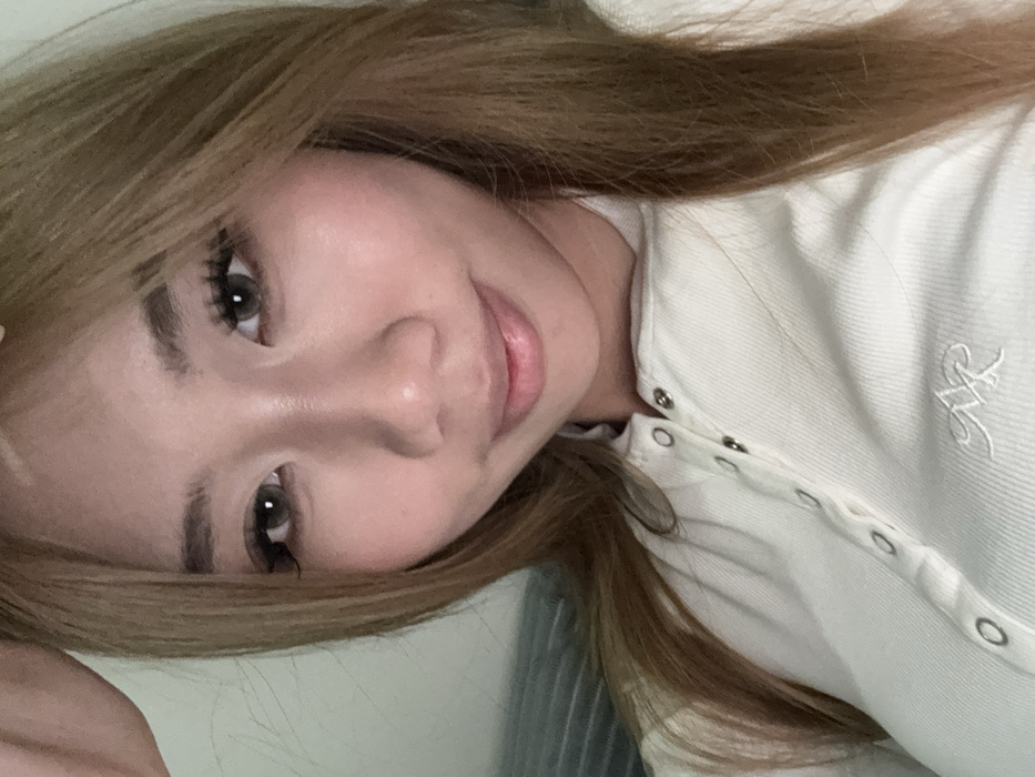 A young East Asian female subject is depicted in a close-up, head-and-shoulders portrait, likely a selfie, within an indoor environment. She possesses long, light brown hair, styled to frame her face, and a fair complexion. Her expression features a subtle smile with closed lips, and she looks directly at the camera. Her eyes appear to have light grey or hazel contact lenses, accentuated by prominent eyelashes and defined eyebrows. A portion of her right hand is visible in the upper left corner of the frame, near her head.
She is dressed in a light-colored, possibly cream or off-white, ribbed long-sleeved top. The garment has a Henley-style placket secured by several silver-colored snap buttons. An embroidered logo, consisting of two stylized, intertwined letters (resembling 'AR' or 'MR'), is visible on the left chest area of her top.
The background is indistinct and out of focus. To the right, a light-colored wall or surface is visible. To the left, a darker, horizontally striped fabric, possibly a blanket or cushion, is partially visible, indicating a typical indoor setting. The lighting is soft and appears to be artificial or diffused natural light. No specific visual cues are present within the image to identify the location as Yuzhong District, China; the environment is generic and consistent with a private indoor residence.