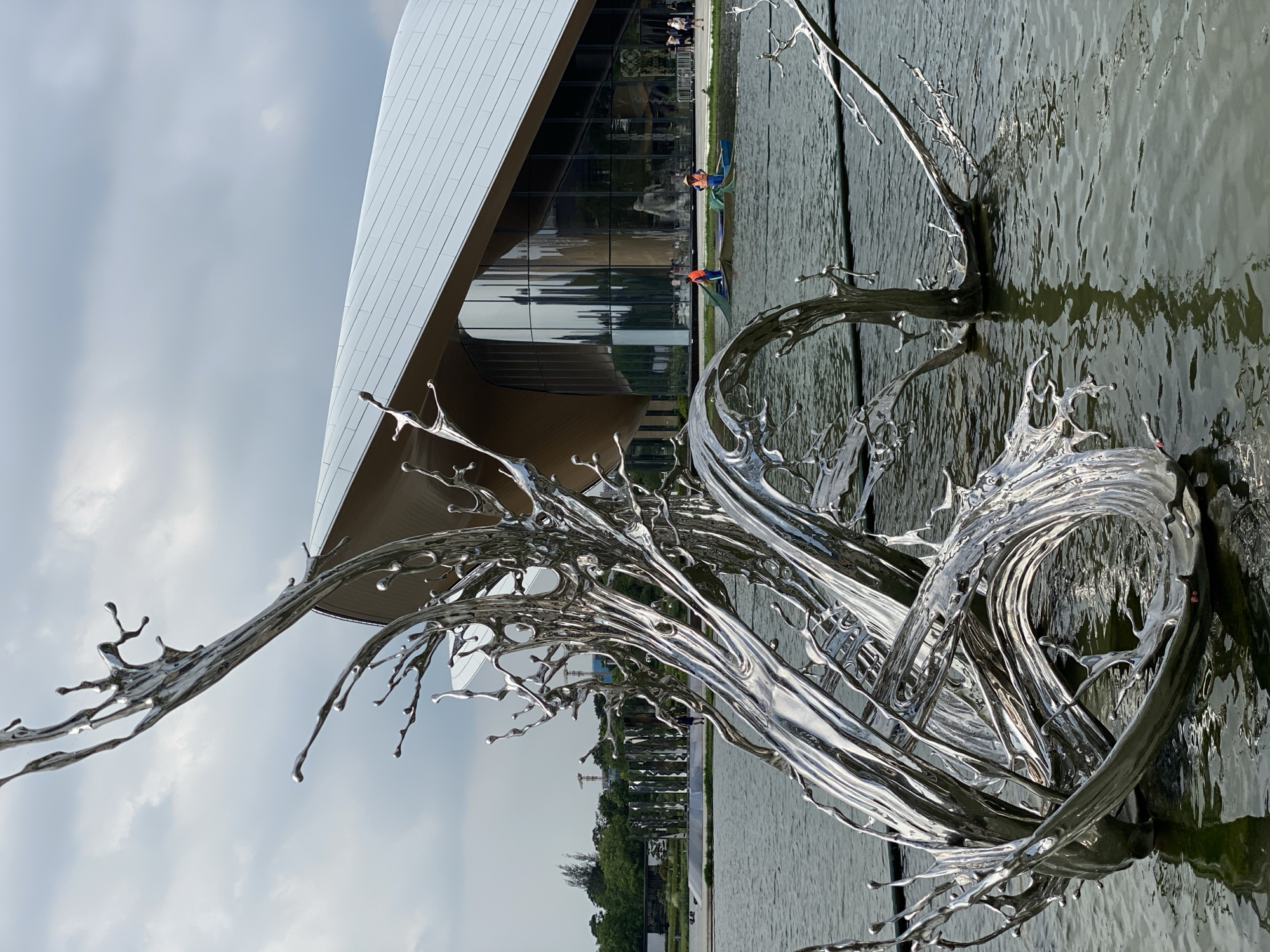 A large, abstract, silver sculpture resembling a water splash is situated in a body of water. The sculpture's metallic surface reflects the cloudy sky and surrounding environment. In the background, a modern building with a white, angled roof and large glass windows is visible. Two people in small boats, wearing orange life vests and hats, are in the water near the building, appearing to be tending to nets. Further in the distance, there are trees and other structures, including what appears to be a cylindrical art installation. The overall setting suggests an outdoor public space, likely a park or waterfront area within a city. The location is identified as Jinniu District, China.