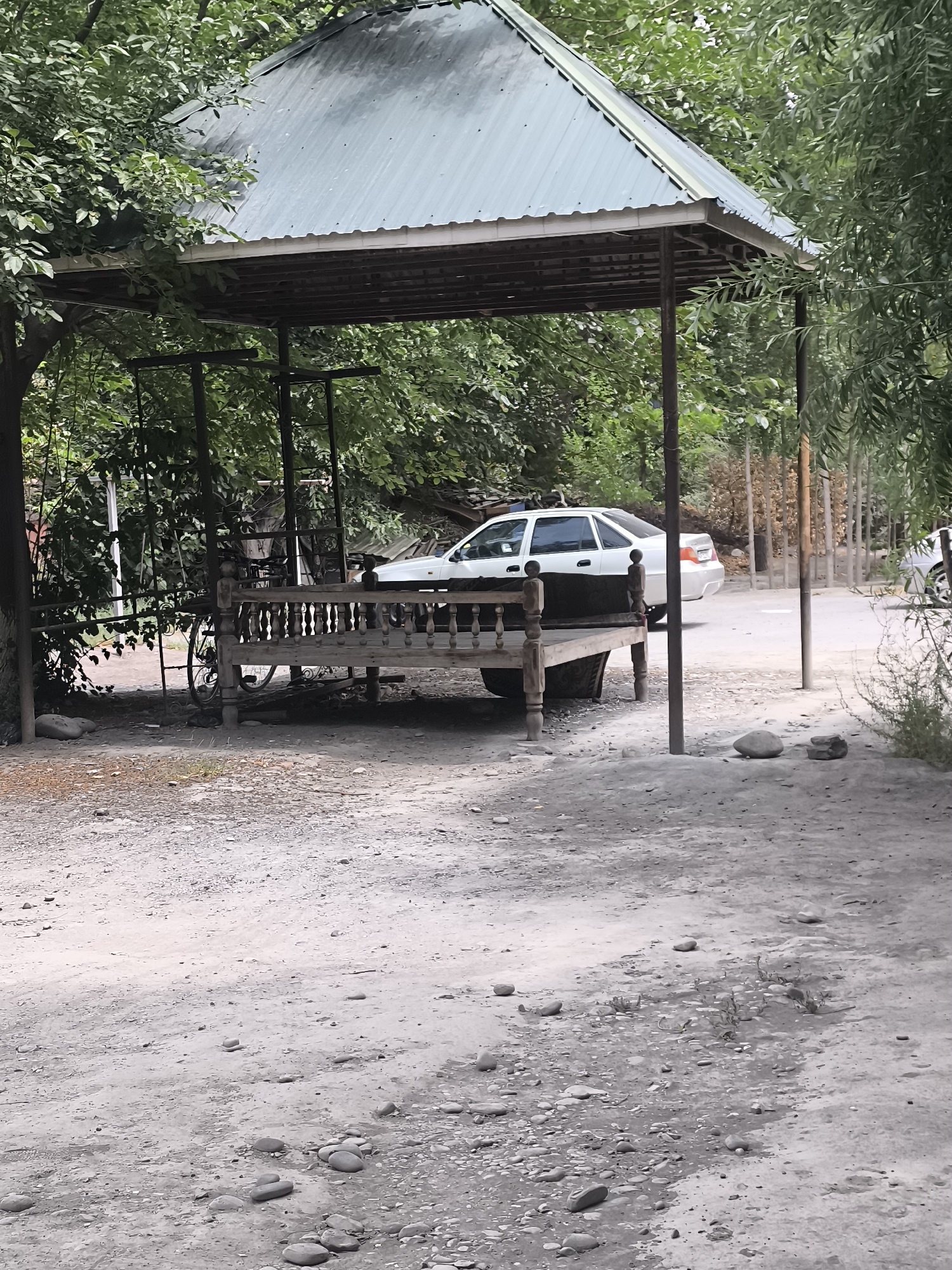 The image shows an outdoor scene in Kokand, Uzbekistan, featuring a covered seating area with a corrugated metal roof supported by metal poles. Beneath the shelter is a wooden daybed or bench with an ornate railing and wooden posts, resting on tires. A bicycle is partially visible behind the daybed. To the right of the shelter, a white sedan is parked on a road, with a light-colored gravel ground in the foreground and dirt and small stones. Lush green trees surround the area, with some wooden posts and fencing visible in the background. The overall impression is that of a rustic outdoor space, possibly a resting area in a garden or courtyard.