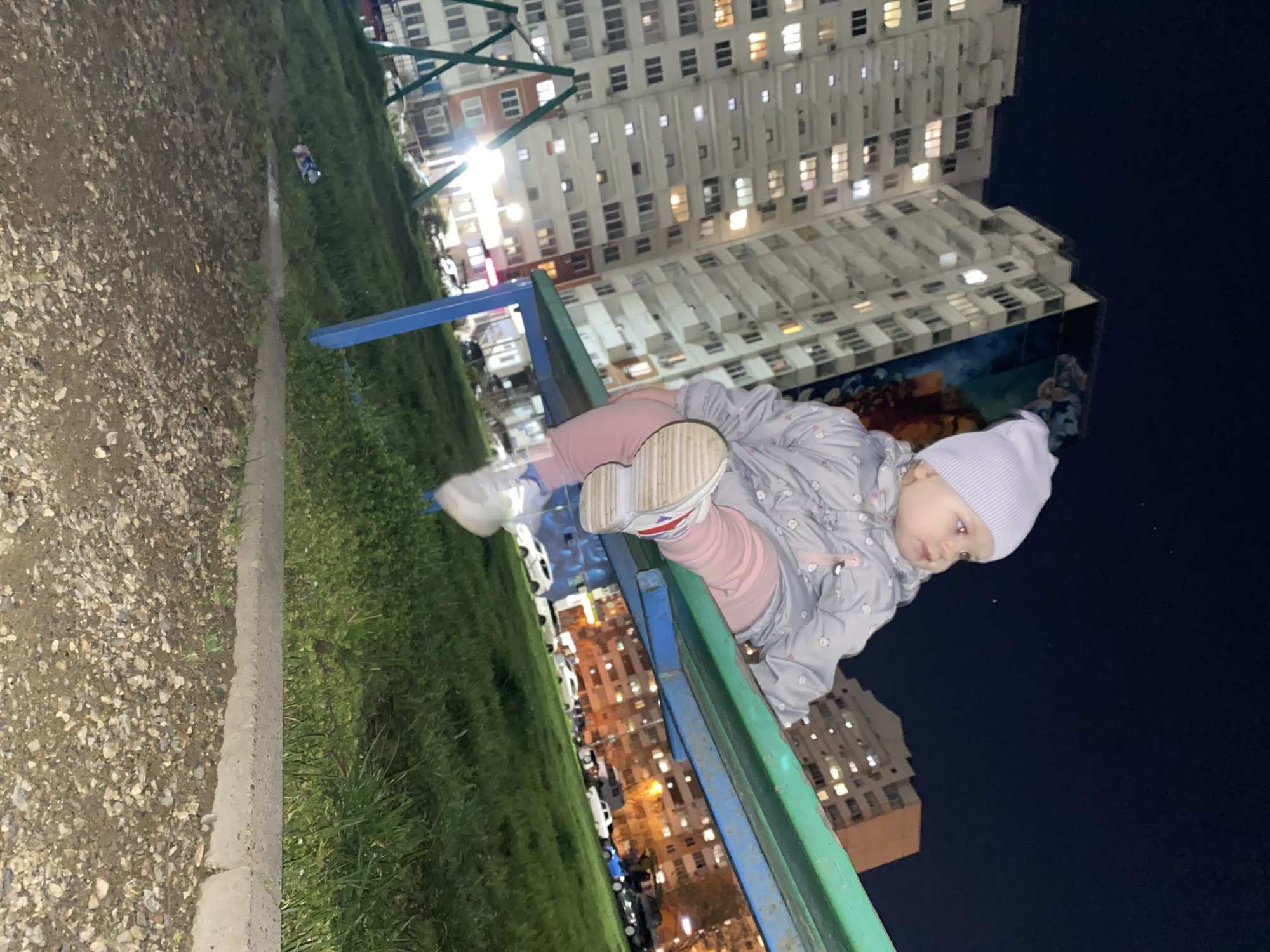 A medium shot depicts a toddler in a gray patterned coat, pink pants, a white hat, and white sneakers sitting on a blue and green park bench at night. The toddler is looking down and to the right. Behind the bench is a grassy area with a sidewalk, followed by a gravel area. In the background, there are several high-rise buildings with many lit windows, indicating nighttime in an urban setting. There is a dark sky in the upper-right corner of the image.