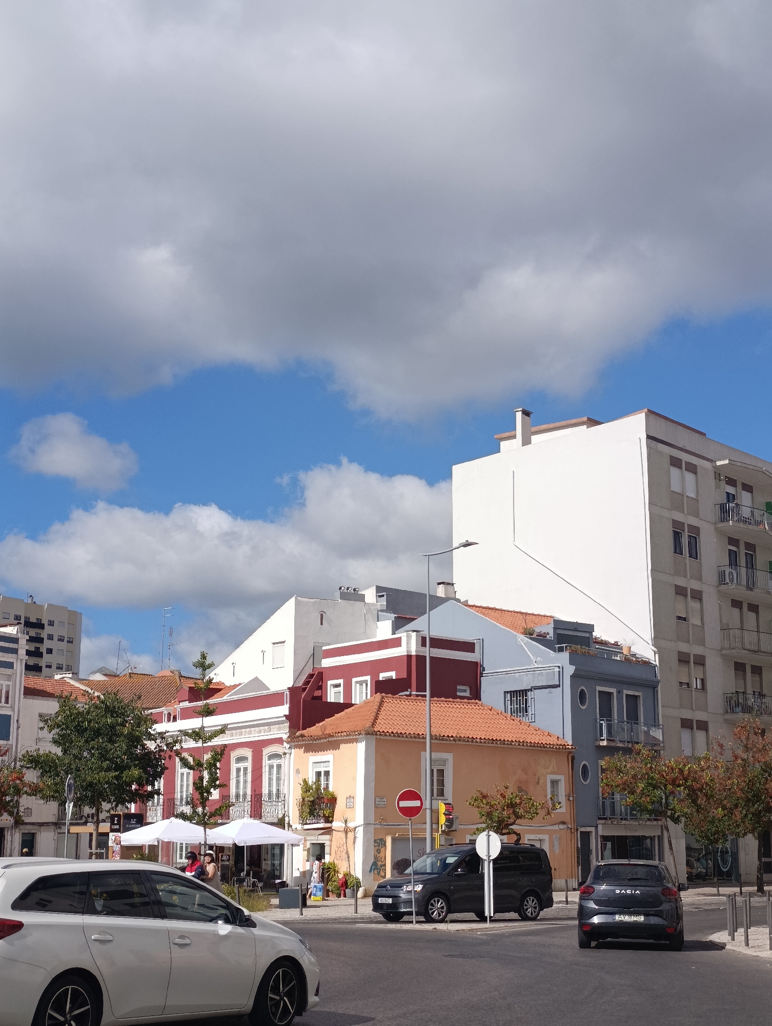 This image depicts a street scene in Setúbal, Portugal, likely in the vicinity of Praça do Bocage, given the architecture. The foreground features a white station wagon driving on an asphalt road. To the right, a dark-colored sedan is parked on the side of the road. In the midground, a row of colorful buildings lines the street. The building on the far right is a multi-story apartment building with balconies. Adjacent to it is a taller, modern white building. Further to the left, there are shorter buildings with red and orange facades, some featuring white trim and balconies. Two white umbrellas are set up outside a cafe or restaurant, with patrons seated beneath them. Trees with autumnal foliage are visible along the street. The sky is blue with scattered white and gray clouds. A street light and traffic signals are also present.