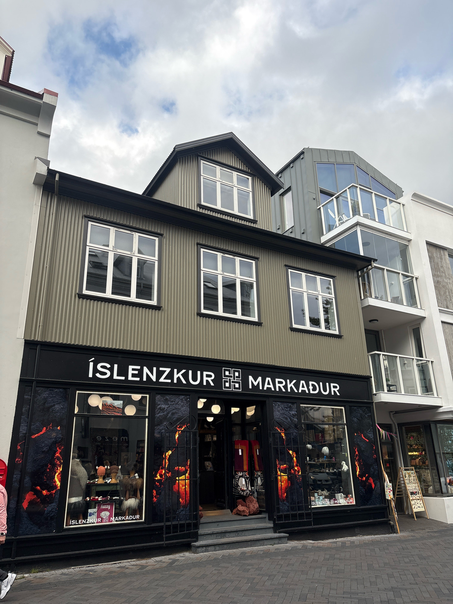 This image shows the storefront of a shop named "ÍSLENZKUR MARKADUR" located on a street in Reykjavik, Iceland. The shop features a black facade with large display windows showcasing knitted goods, hats, and other merchandise. The windows are decorated with a fiery graphic design. The building itself is multi-story, with the upper floors featuring traditional Icelandic architecture and modern additions. Adjacent to the shop is a contemporary building with glass balconies. The street is paved with cobblestones, and the sky above is overcast. There are a few people visible in the foreground and on the street.