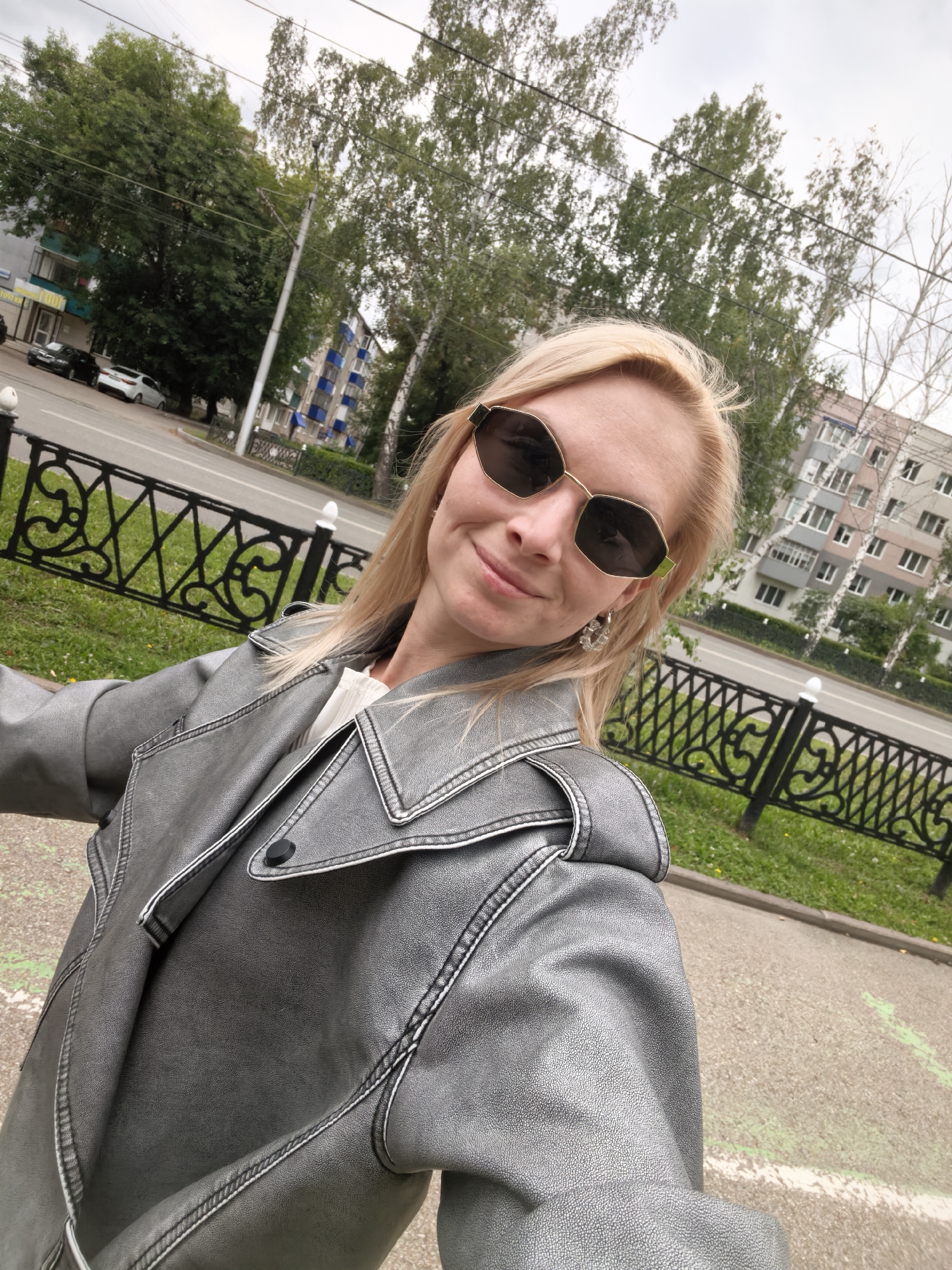 A blond woman wearing sunglasses and a grey jacket is taking a selfie outdoors in an urban environment. She is standing on a sidewalk with a black metal fence to her left, separating her from a grassy area. Beyond the grass is a street with cars and residential buildings. The buildings appear to be multi-story apartment blocks. Trees are visible behind the buildings, and the sky is overcast. The woman is smiling, and her blonde hair is blowing slightly in the wind. The image appears to be taken in Sterlitamak, Russia, given the architectural style of the buildings and the overall urban landscape.