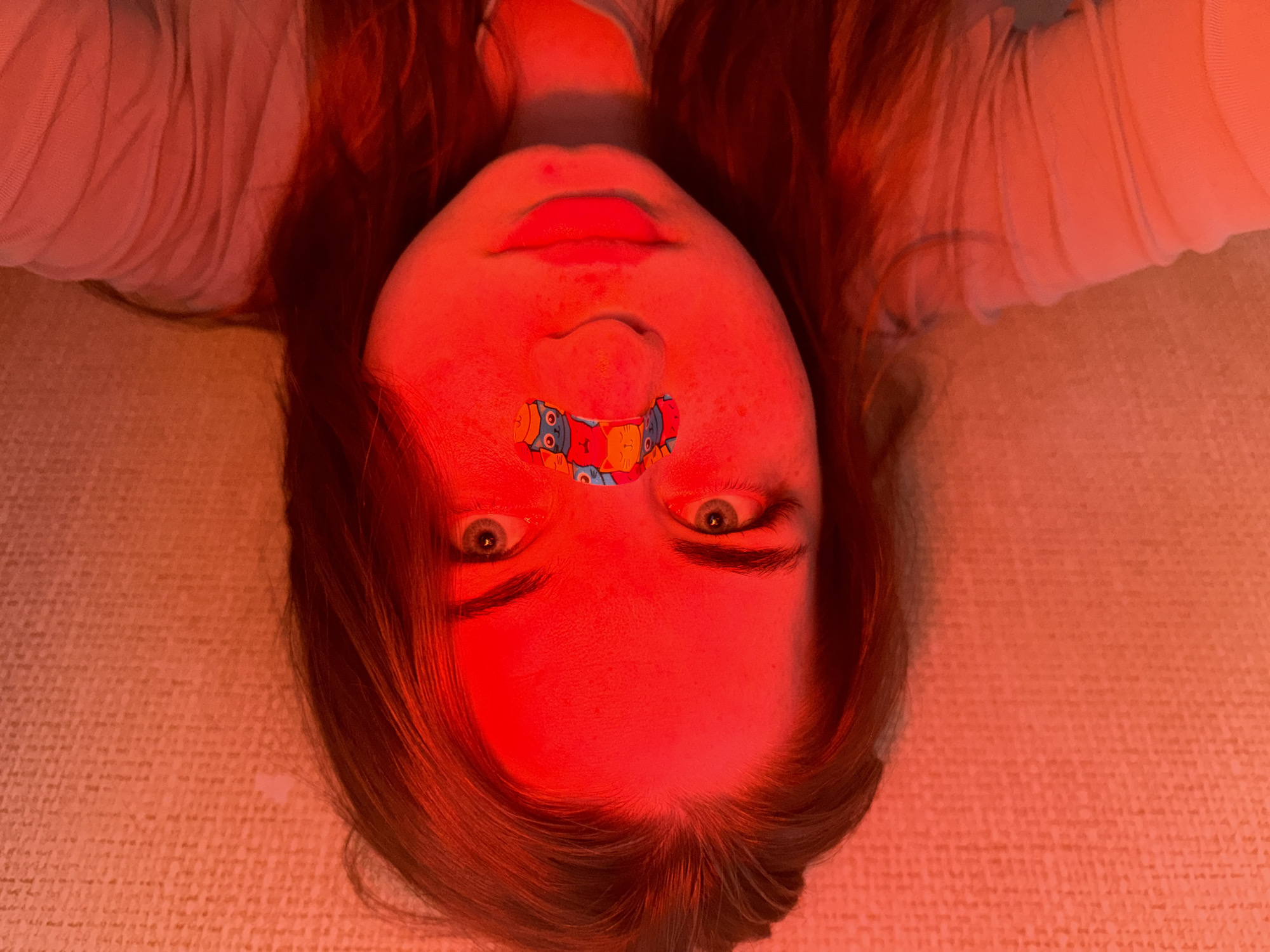 A close-up, upside-down view shows a fair-skinned person lying on a textured surface, possibly a bed or couch. The subject has reddish-brown hair and is wearing a light-colored garment, possibly a shirt. A brightly colored bandage with cartoon cat and owl designs is affixed to the area between their nose and upper lip. The face is illuminated with a strong red light source, obscuring fine details but revealing the person's eyes, which are open and gazing at the camera. The location is unknown; there is no indication that the image was taken in Chaikovsky, Russia.
