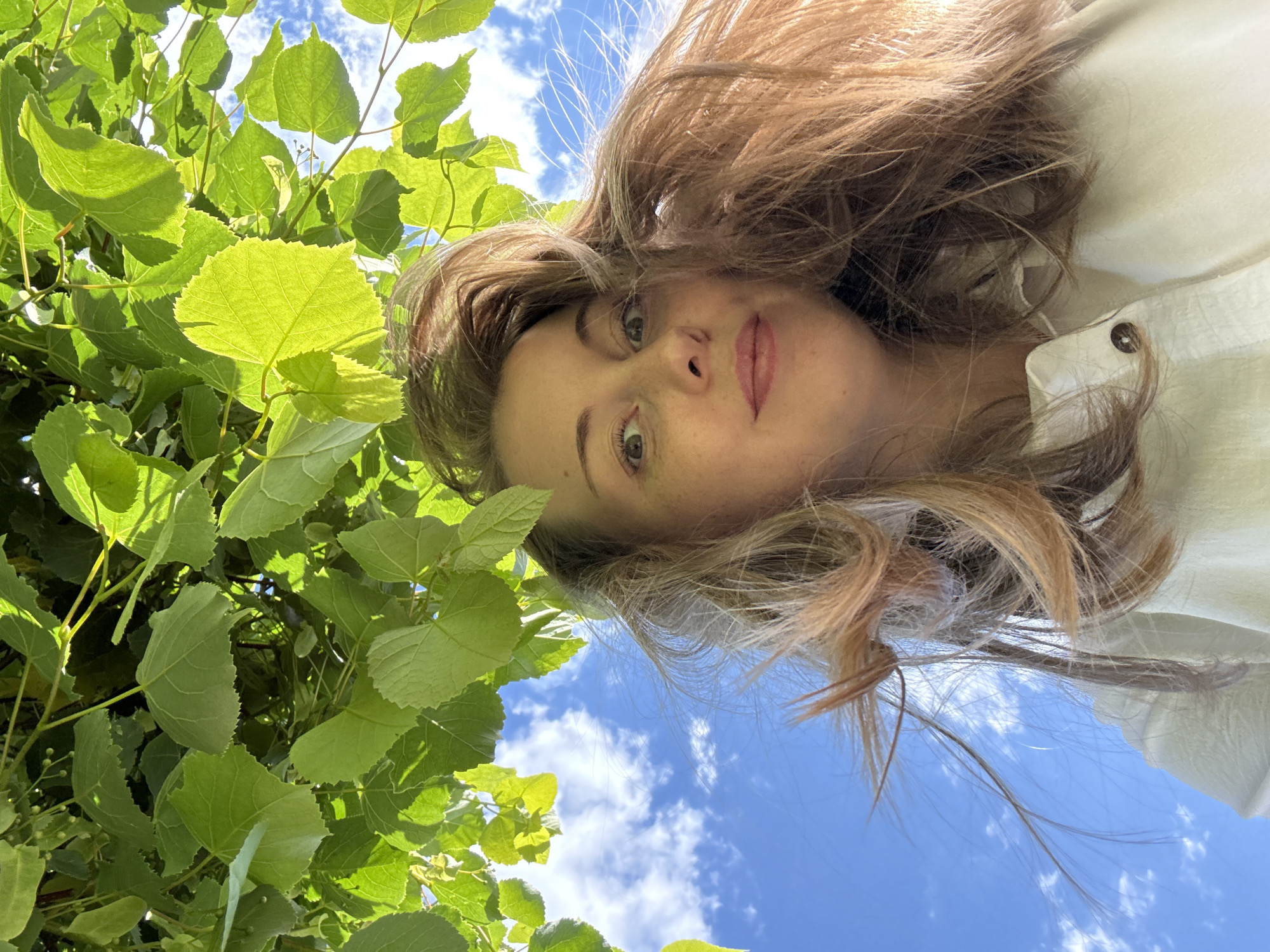 The image captures a low-angle upward-facing selfie of an adult female positioned beneath a dense canopy of green leaves and a blue sky with scattered white clouds. The woman, with light-colored skin and reddish-brown, windswept hair, occupies the lower portion of the frame. She has visible freckles across her nose, and her eyes are wide with a slightly surprised or curious expression; her lips are slightly parted. She is wearing a light-colored, possibly white, collared shirt, featuring a single brown button visible on the placket. The upper half of the image is dominated by vibrant green, serrated-edge leaves, likely from a deciduous tree such as a linden, which are backlit by bright sunlight, casting varied shadows and highlights. Small green buds or immature fruits are discernable among the foliage. Patches of clear blue sky with sparse white cumulus clouds are visible through the leaves and to the left of the woman's head. The specific location is Unknown, Unknown.