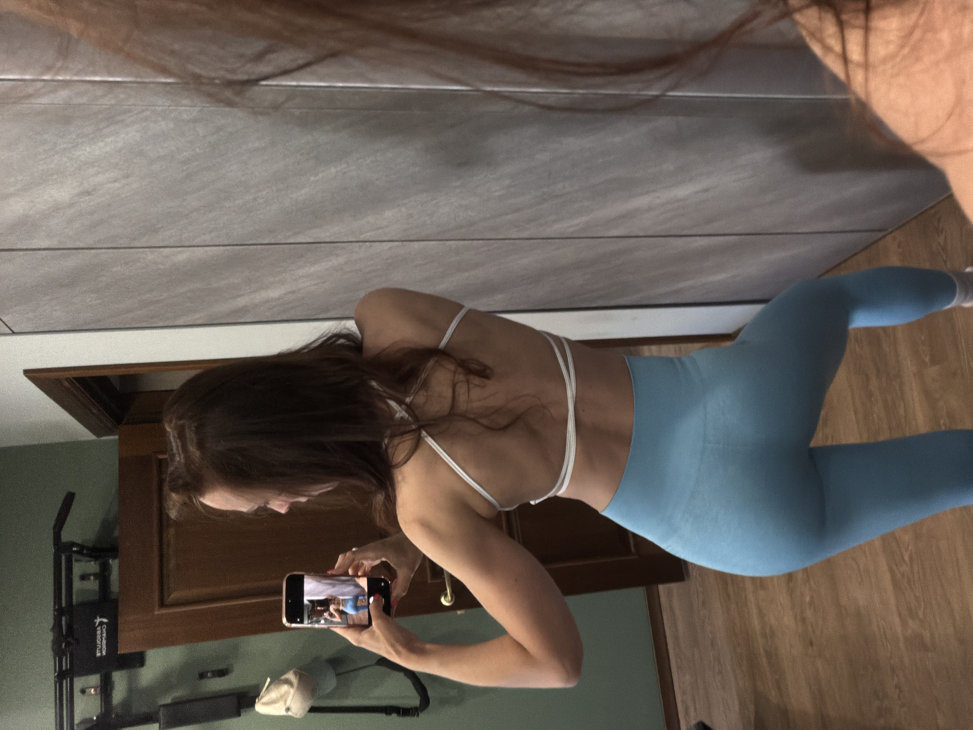 The image, rotated 90 degrees counter-clockwise, shows a woman taking a mirror selfie indoors. The woman has long brown hair and is depicted from her back and right side, wearing a white sports bra with multiple thin straps and light blue high-waisted athletic leggings. She is holding a rose gold smartphone in her right hand, pointed towards a reflective surface. The screen of the phone clearly displays her own reflection, confirming she is capturing a selfie.
The setting appears to be a home gym or an exercise area within a room. The primary background feature is a large wall covered with grey, rectangular tiles, which serves as the reflective surface for the selfie. To the right of the woman (when the image is upright), a light brown wooden floor is visible. Behind her, there is a dark brown wooden doorframe and a dark green painted wall. Against this green wall, a black piece of fitness equipment, resembling a pull-up bar or power tower, is mounted. A light-colored baseball cap hangs on or near the equipment.
Notable details include the woman's red-painted fingernails. On the fitness equipment, partially visible text can be seen, which includes the word "ABSOLUTELY" along with other less discernible words. The lighting in the scene appears to be indoor artificial light. No specific time of day or outdoor weather conditions can be determined from the image.
