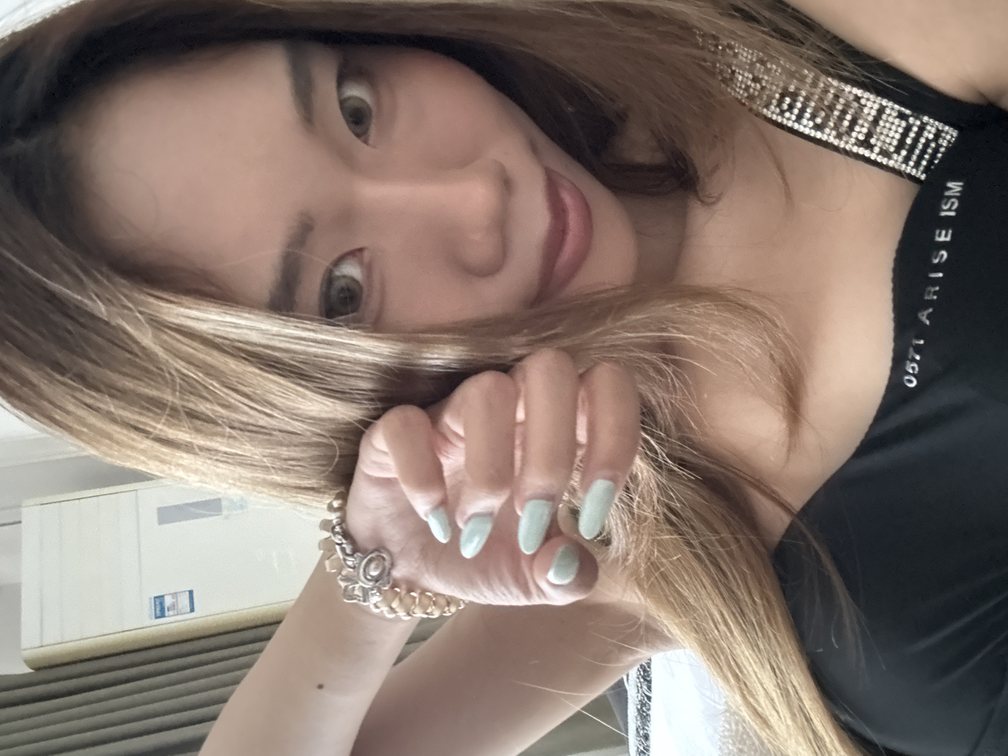 A close-up, eye-level shot shows a young East Asian woman with long, light brown hair and fair skin, posing indoors. She is wearing a black top with a jeweled strap and text that reads "0571 ARISE ISM" on the hem. Her left arm is raised, with her hand near her face, showcasing light blue painted fingernails and a silver bracelet. The background includes a beige air conditioner unit and a grey curtain. The lighting is soft and natural.