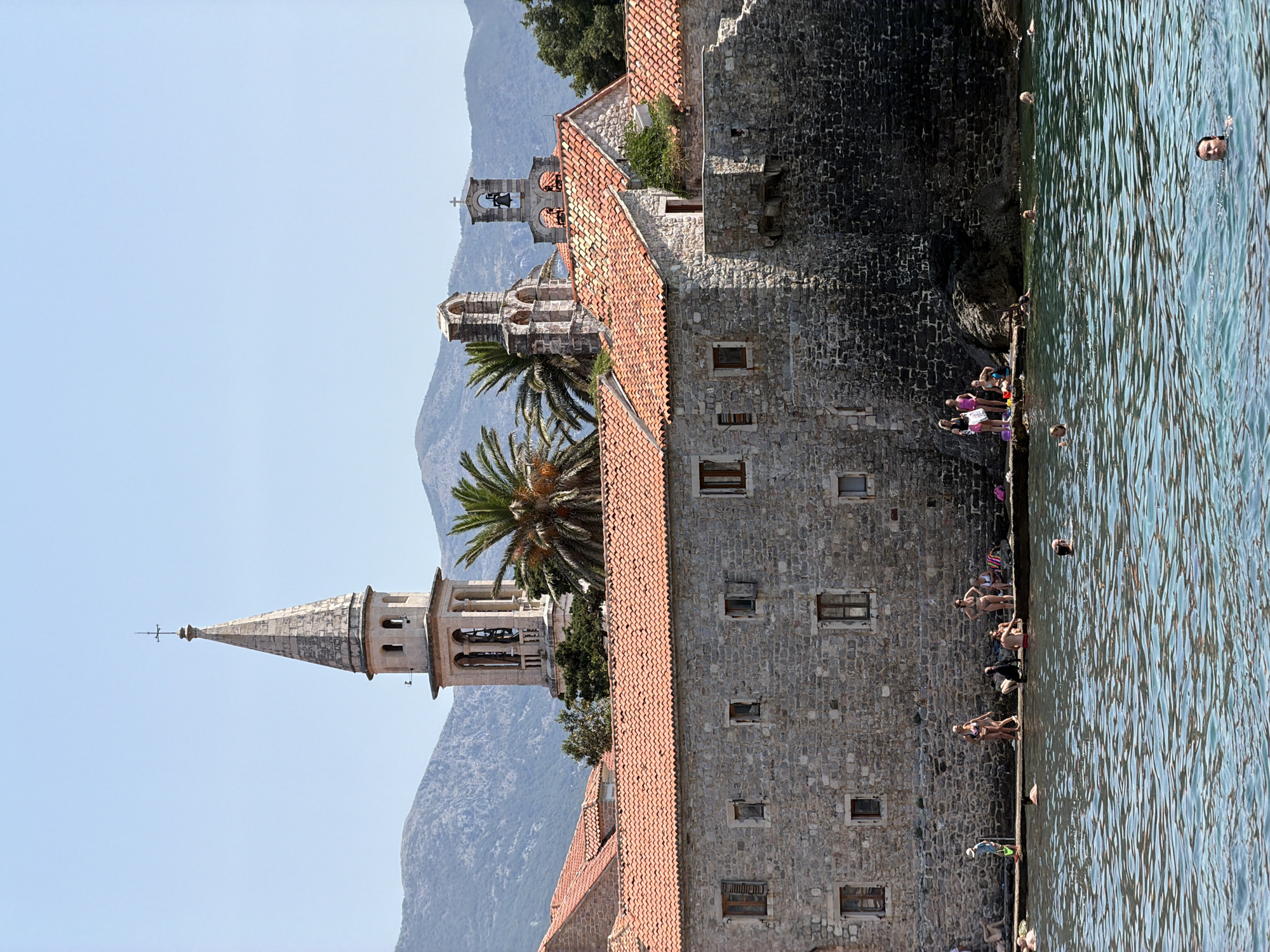 This is an eye-level, medium shot of a seaside view of Budva, Montenegro, featuring historical stone buildings with red tile roofs and a bell tower in the background. The water in the foreground is a clear blue, with several people swimming and wading. Along the stone wall of the buildings, a group of people are gathered, some sitting and others standing. Palm trees are visible behind the buildings. The sky is a clear, bright blue, and distant mountains are visible.