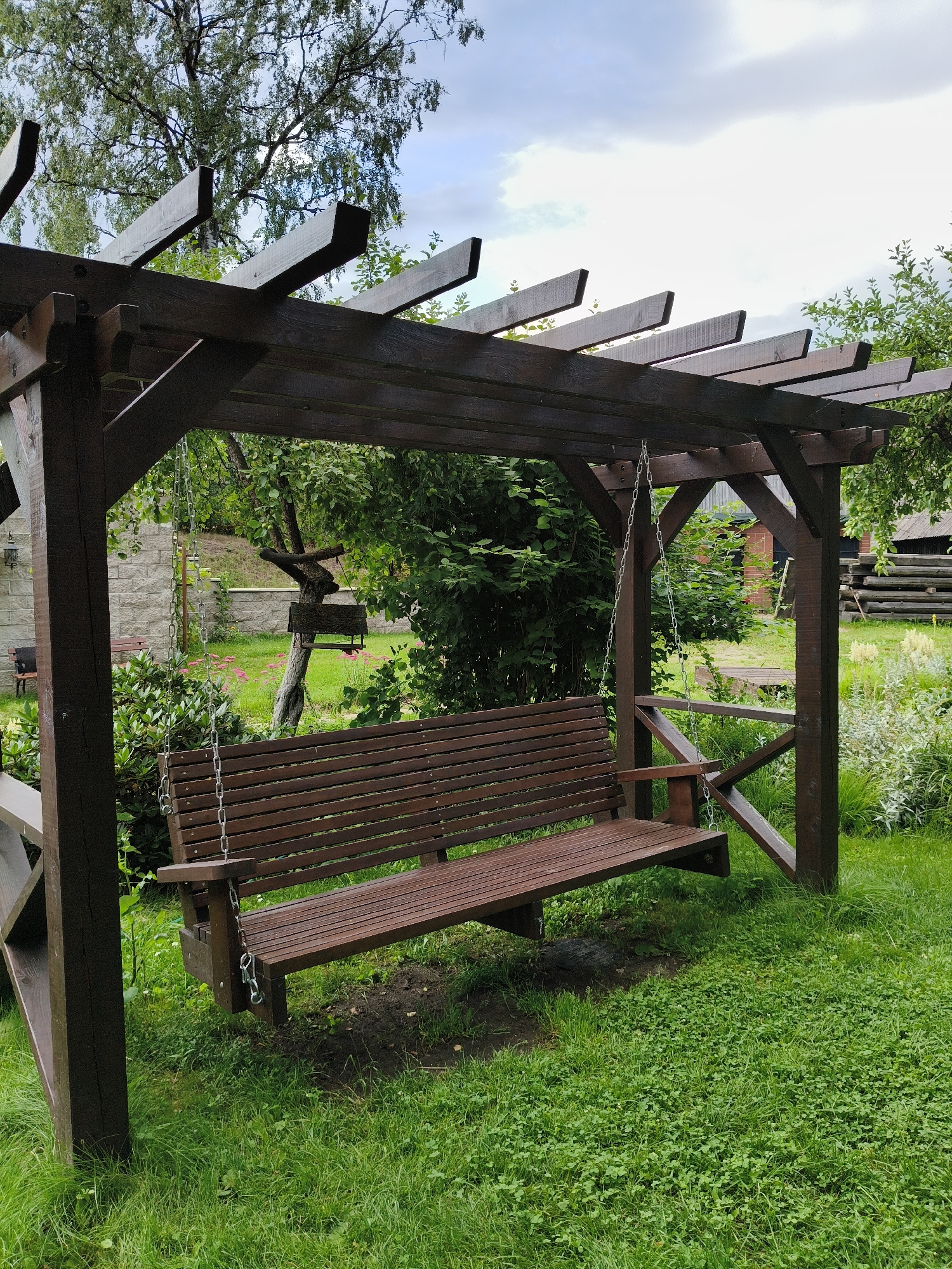 A wooden swing set with a bench is located in a grassy yard. The swing set is dark brown and has a slatted roof. The bench is also dark brown and has a slatted seat and back. In the background, there are trees, bushes, and a stone wall. A cloudy sky is visible above the trees. The scene appears to be in a rural or suburban setting.