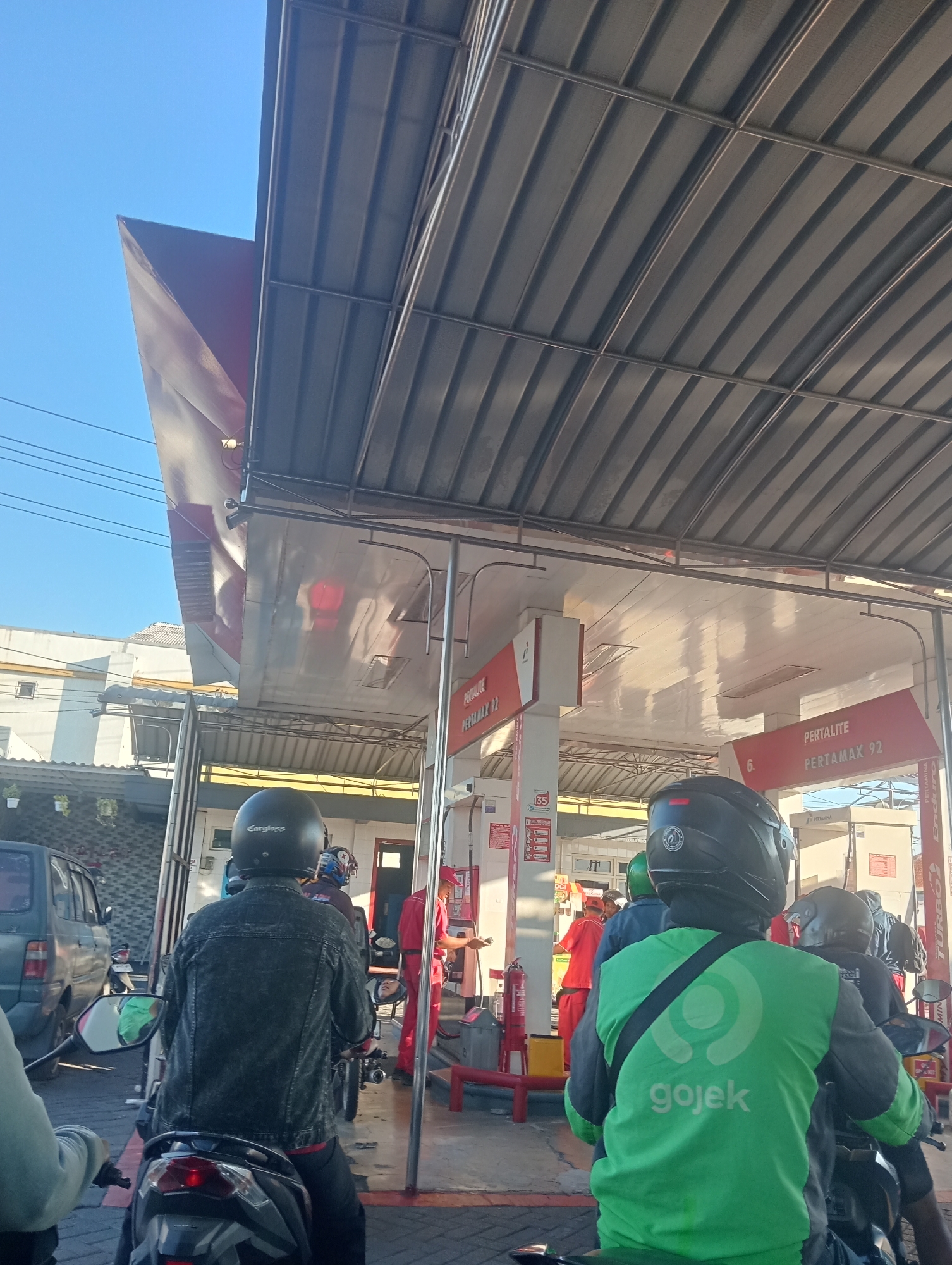 In Sawotratap, Indonesia, this image captures a queue of motorcyclists at a Pertamina gas station under a corrugated metal roof. The perspective is from behind a rider wearing a green Gojek jacket and a black helmet. To the left, a dark green car is parked, and further back, a building with white siding and a sloped red roof is visible. The gas station features red and white signage indicating "PERTAMAX 92" and "PERTALITE". In the background, two workers in red uniforms are attending to customers. The sky is clear and blue.