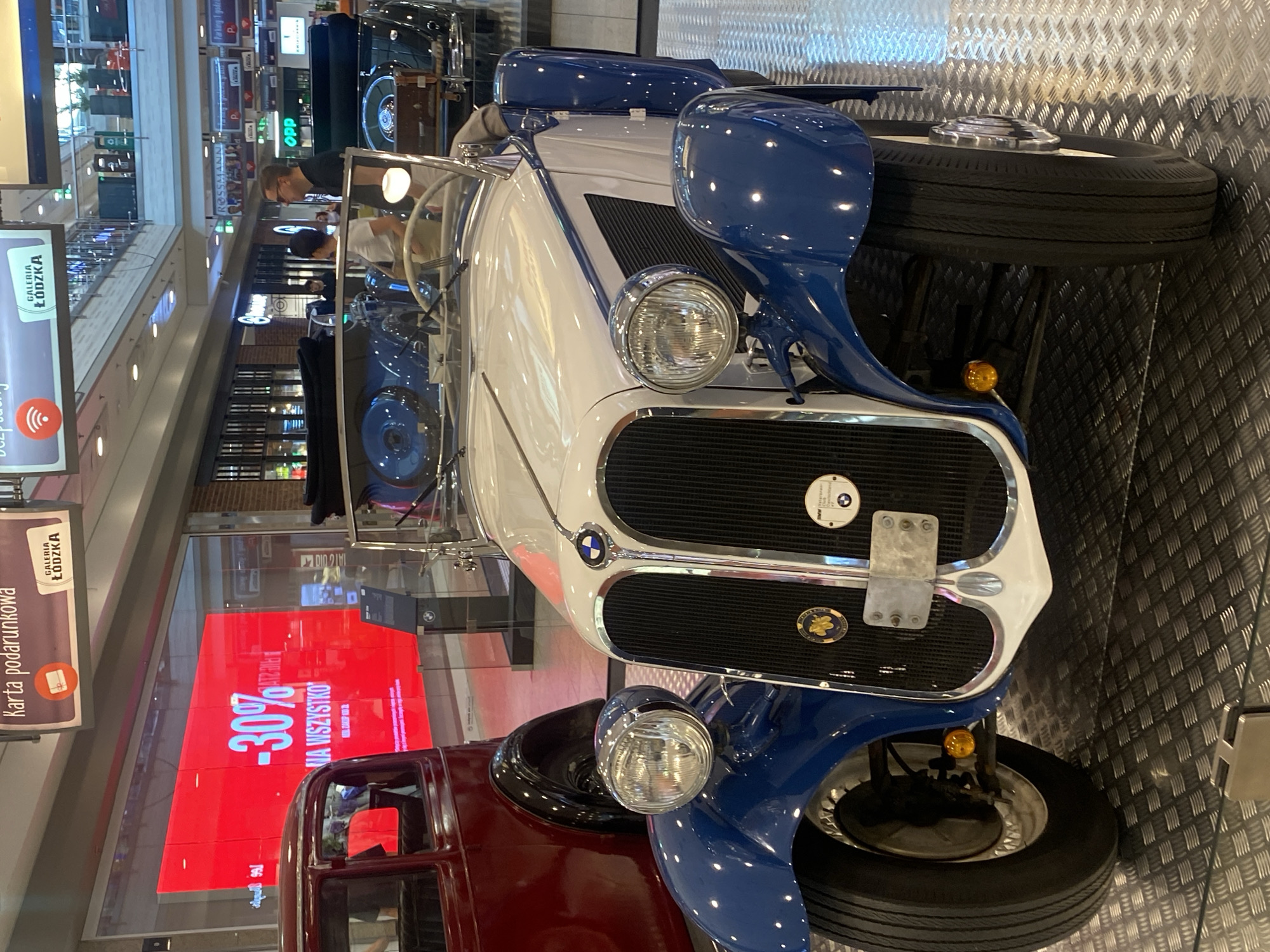 This image captures a display of vintage cars, likely in a shopping mall or exhibition space, as suggested by the signs for "Galeria Łódzka" and retail stores like "Rossmann." The primary focus is a classic white and blue convertible, which is positioned prominently in the foreground. Its chrome accents, round headlights, and distinctive grille are clearly visible. To the left, a portion of a dark red vintage car is also visible.
In the background, several people are present, seemingly observing the cars. One individual, a man wearing glasses and a dark t-shirt, is looking at the convertible. Another person, wearing a white top, is also near the car. The setting appears to be indoors, with the cars displayed on a textured floor.
A large red digital screen in the background displays promotional text, with " -30% NA WSZYSTKO" (30% OFF EVERYTHING) being the most legible part. Other signs in Polish are visible, including "Karta podarunkowa" (Gift card) and "Galeria Łódzka," confirming the location in Łódź, Poland. The overall atmosphere suggests a public event or exhibition showcasing automotive history. The lighting is artificial, characteristic of an indoor environment.
