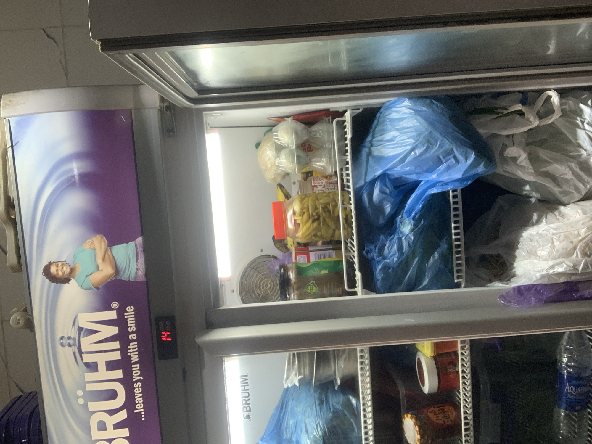 This is a close-up, eye-level, indoor shot of an open commercial refrigerator, likely in a food service establishment in Abuja, Nigeria. The refrigerator is filled with various food items and packaging. The top portion of the refrigerator displays a large advertisement for "BRÜHM" appliances, featuring a smiling woman and the tagline "...leaves you with a smile." A digital temperature display shows "14" degrees Celsius.
Inside the refrigerator, the shelves are filled with items primarily in blue and white plastic bags, suggesting preserved or prepared food. Visible items include what appear to be jars of pickles or preserved vegetables, round containers, and possibly raw ingredients. On the right side, a clear plastic container holds what looks like eggs. The overall impression is of a busy, functional kitchen or storage area.