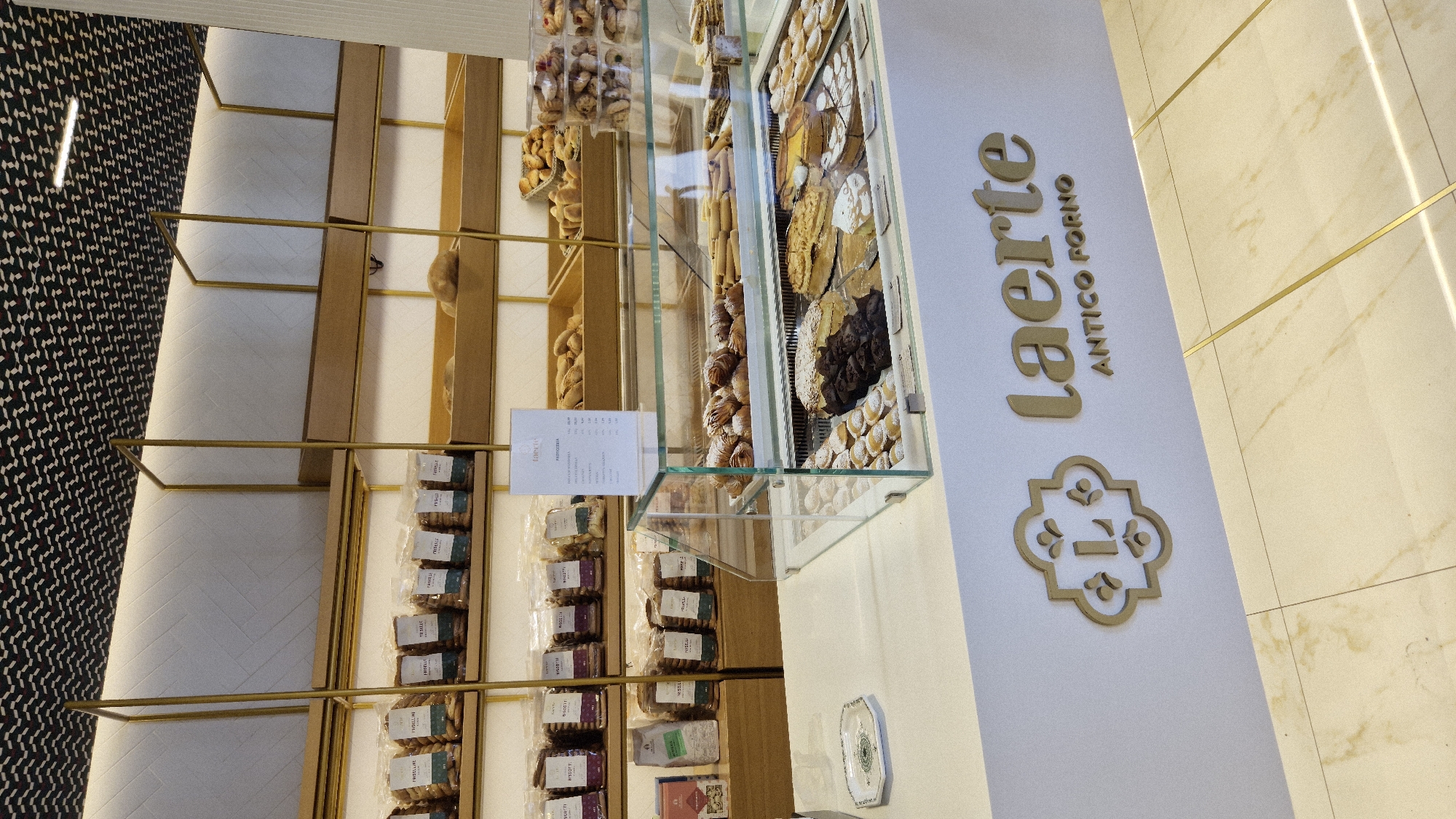 The image shows the interior of a bakery, likely in Laterza, Italy, given the text on the counter. The bakery features modern, clean aesthetics with white tiled walls and shelves. Gold accents are present in the shelving units and on the store's logo.
In the foreground, a white counter displays a glass-enclosed refrigerated case filled with various pastries and baked goods. These include small powdered sugar cookies, chocolate brownies, a large cake with cream and fruit, and tarts. Behind the counter, shelves are stocked with packaged cookies and other baked items, some of which are branded "Friselline." A small white sign with text in Italian lists pastry options and prices.
Above the shelves, a patterned dark ceiling design adds a contemporary touch. The floor appears to be tiled with a marble-like pattern. The store's logo, a stylized "L" within a decorative frame, is visible on the front of the counter, along with the name "laerte" and "ANTICO FORNO" below it.
There are no people visible in the image, and the scene is static, depicting a well-organized retail environment. The lighting is bright and even, illuminating the products and the interior design.