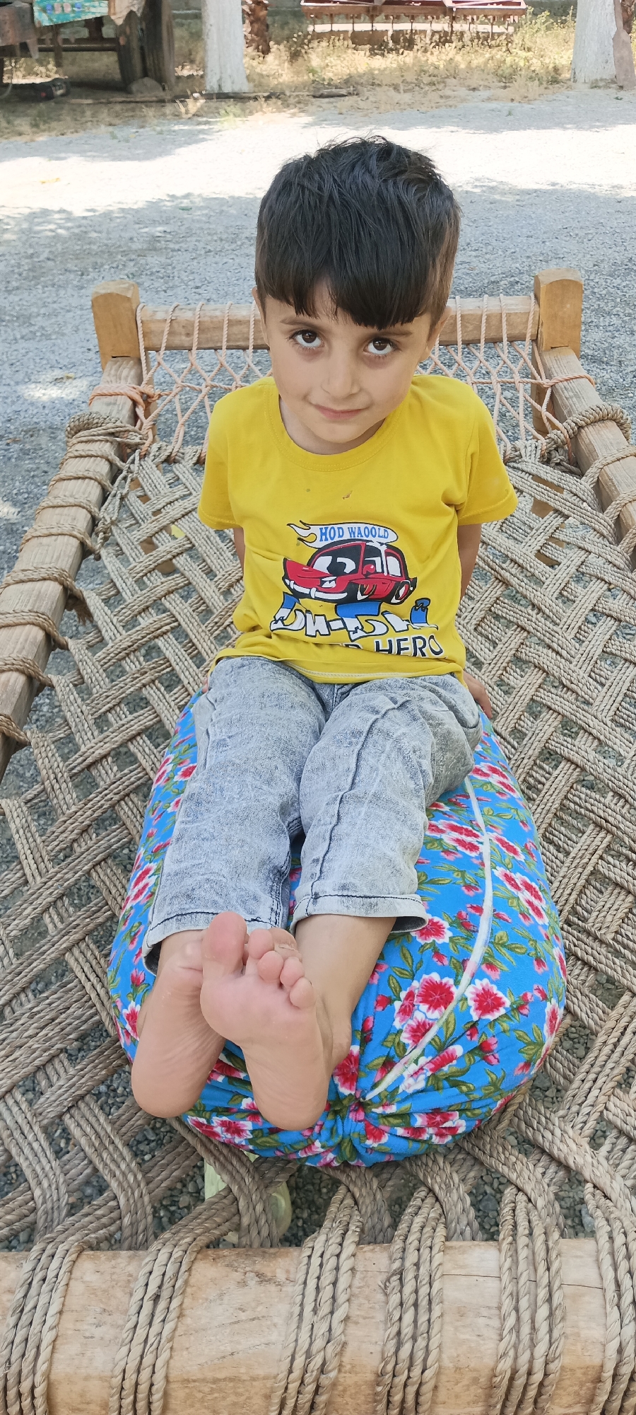 A young child sits cross-legged on a floral-patterned cushion atop a woven cot. The cot, constructed from wooden beams and woven rope, is set outdoors in what appears to be Alam Khan Kalay, Pakistan, judging by the landscape, gravel ground, and simple construction. The child wears a yellow T-shirt with a cartoon car graphic and light-wash jeans. The child's bare feet are visible and face toward the viewer. The background contains a light scattering of vegetation and an old wooden wagon.