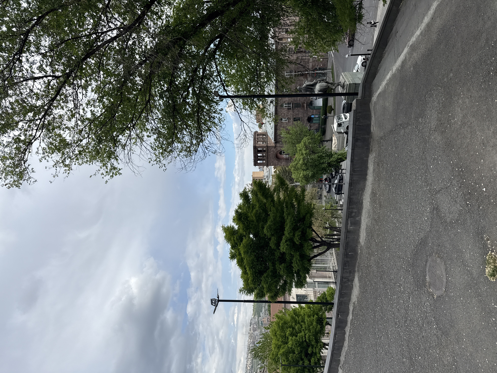 The image is an eye-level, outdoor shot taken in Yerevan, Armenia, showcasing a blend of urban and natural elements.
Dominating the upper left and center of the frame is a view into the sky where light grey clouds and patches of blue sky are visible.
Along the right side of the image, at street level, is an asphalt sidewalk, containing a manhole. There is a concrete curb marking the edge of the sidewalk, separating it from the road. Beyond the road, there are cars parked.
Further into the background, there is a large stone building with a distinctive bell tower. Trees of various sizes are interspersed throughout the scene. A statue and a public square are visible near the building, providing a glimpse into the city's architecture and public spaces. A lamp post is present in front of the sky.
The presence of trees and the sky suggests the image was taken on the top of a hill.