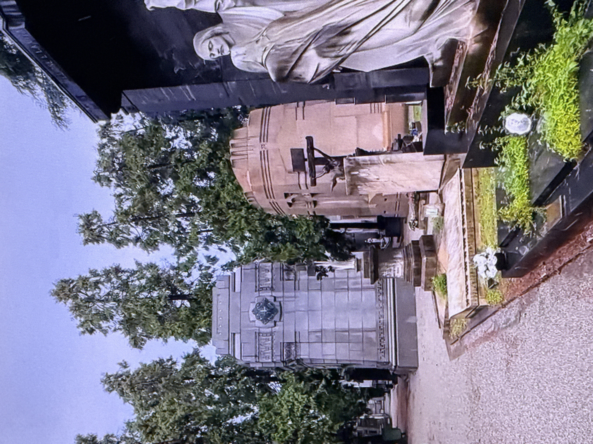 The image depicts a cemetery with large, ornate mausoleums and statues. The sky is overcast, suggesting a cloudy day. Tall, slender trees line the pathways between the structures. In the foreground, a pathway is visible, paved with small, light-colored stones. To the right, a dark, rectangular mausoleum features a large, light-colored statue of a robed figure, possibly Jesus Christ, with one arm raised. In front of this mausoleum, a dark, tiered structure is overgrown with green foliage. Further back, to the left, a large, multi-faceted gray stone mausoleum stands prominently, adorned with a decorative emblem. Several other tombstones and mausoleums are visible in the background, interspersed with trees. The overall impression is one of grandeur and solemnity, typical of an established cemetery. The image does not provide any indication of the location being in Dubai, United Arab Emirates.