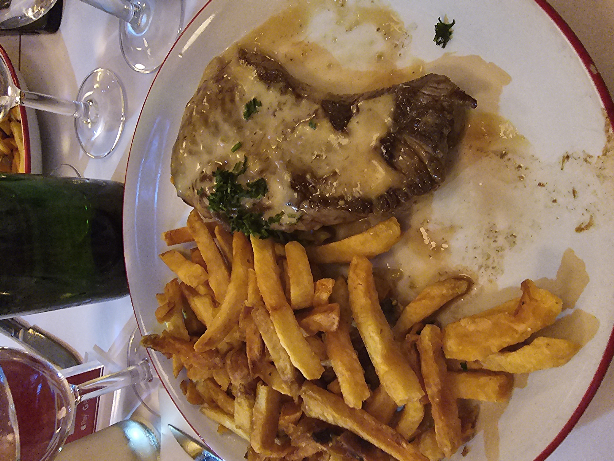 The image shows a partially eaten meal of steak with a white sauce and french fries on a white plate with a red rim, likely in a restaurant in Paris, France. A green bottle and several wine glasses are visible on the table, indicating a dining setting. The plate is partially consumed, suggesting a recent meal. The lighting is artificial, and the composition is from an overhead perspective.