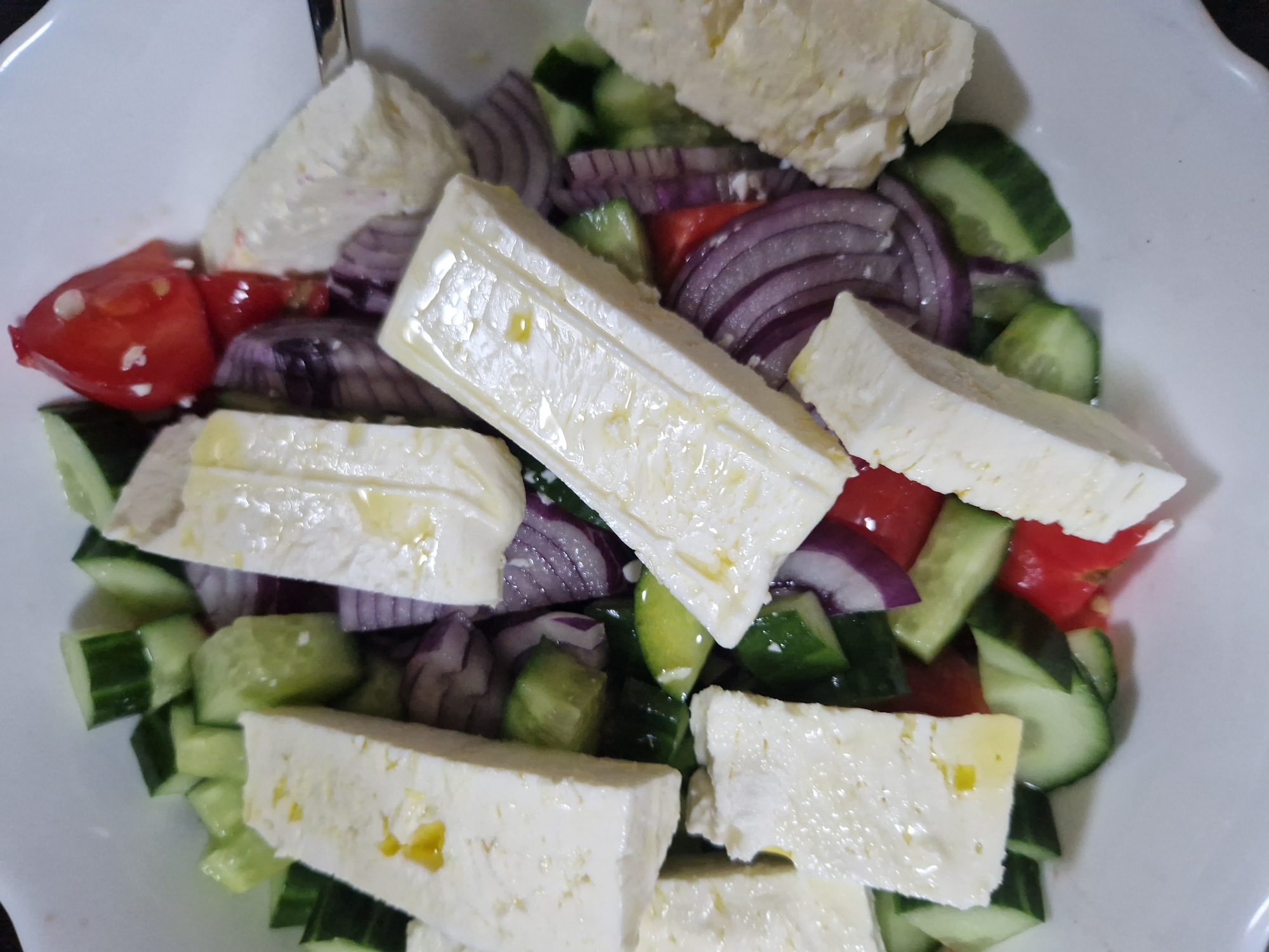 A high-angle, close-up shot of a Greek salad in a white bowl. The salad contains diced cucumbers, red onion slices, chunks of red tomato, and rectangular slabs of feta cheese. Some of the cheese pieces appear to have a light coating of oil. The bowl is most likely in an indoor setting. There is no information available to determine the location in Sofia, Bulgaria.