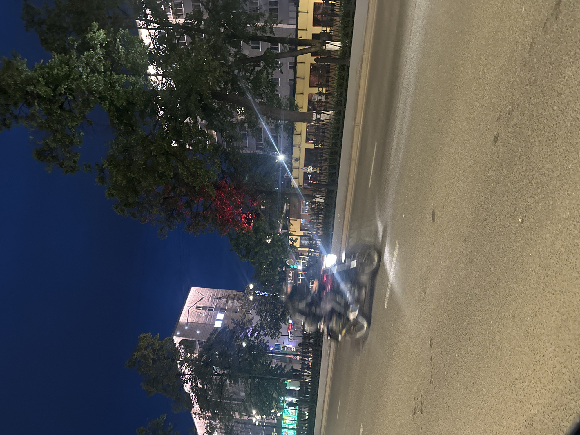 A motorcycle is captured in motion at night on a paved road, with its headlight illuminated. The rider is blurred due to the camera's motion. In the background, multi-story buildings line the street, featuring illuminated windows. Trees with lush foliage are present on the left and right sides of the scene. The sky is a deep, dark blue, suggesting nighttime. The overall environment depicts an urban setting with streetlights and architectural elements characteristic of a city. The location is specified as Perm, Russia.