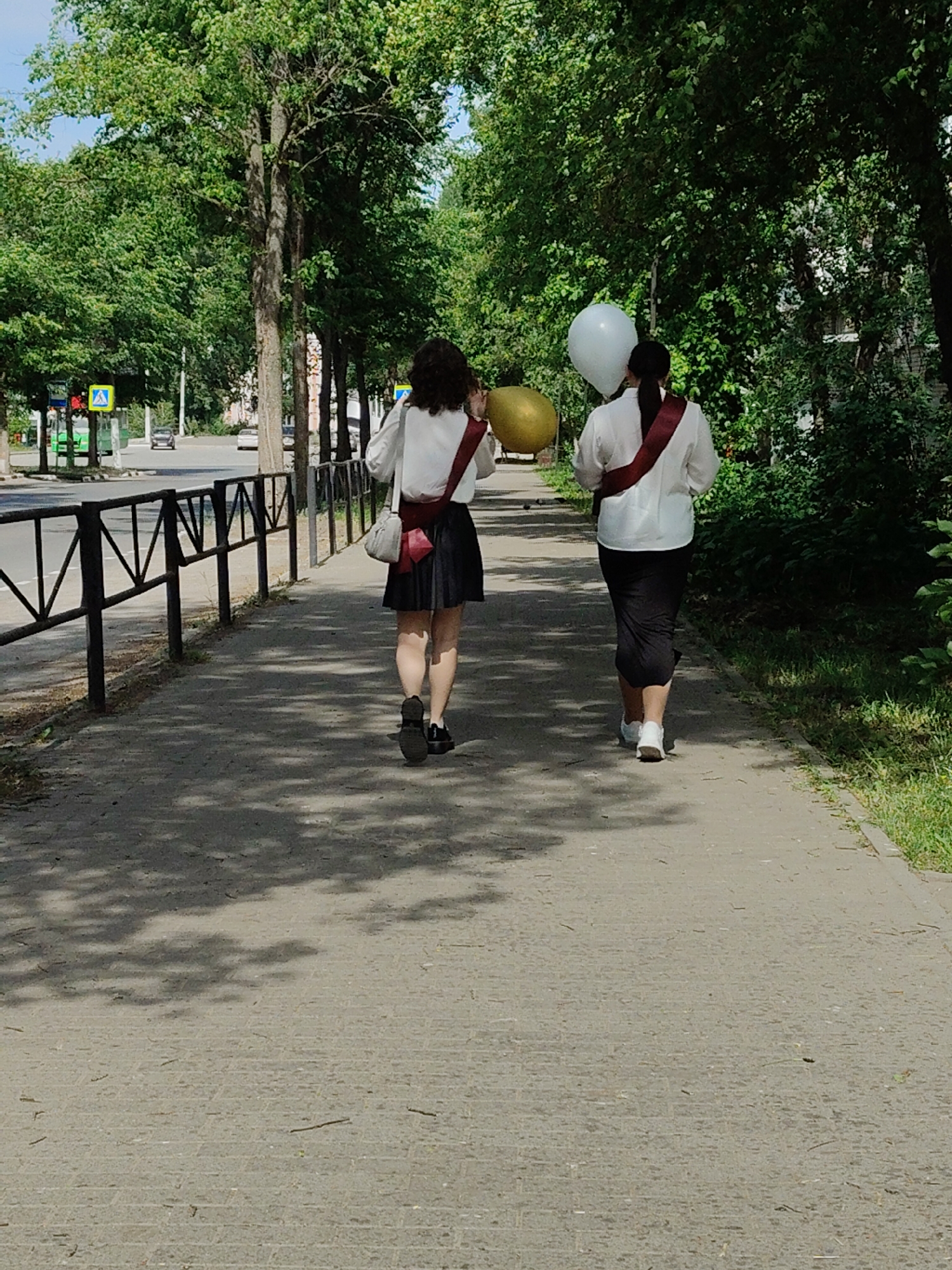Two young women walk away from the viewer on a sidewalk in Unknown, Unknown. The women both wear white shirts and dark skirts, and they have red sashes across their shoulders. The woman on the left carries a gold balloon and a small purse, while the woman on the right carries a white balloon. A black metal fence lines the sidewalk on the left. Trees line both sides of the sidewalk, creating a shaded path. In the background, a road with a crosswalk is visible.