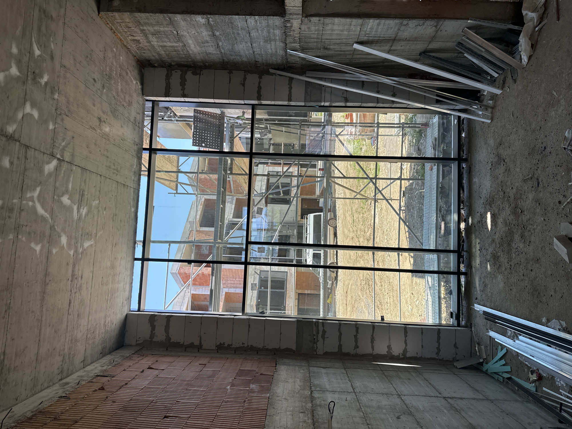 This image depicts the interior of a building under construction in Stara Zagora, Bulgaria. The scene is viewed from within a room, looking out through a large, floor-to-ceiling window. The interior walls are constructed of rough concrete and brick, with some sections appearing unfinished. Debris and construction materials, including metal profiles and blue foam insulation, are scattered on the floor.
The window offers a view of an outdoor construction site. Multiple buildings are in various stages of completion, with visible brickwork and scaffolding. A white van is parked on the dirt ground outside. The sky is clear and blue. The lighting suggests it is daytime. The image appears to be a still photograph taken from a fixed position.
