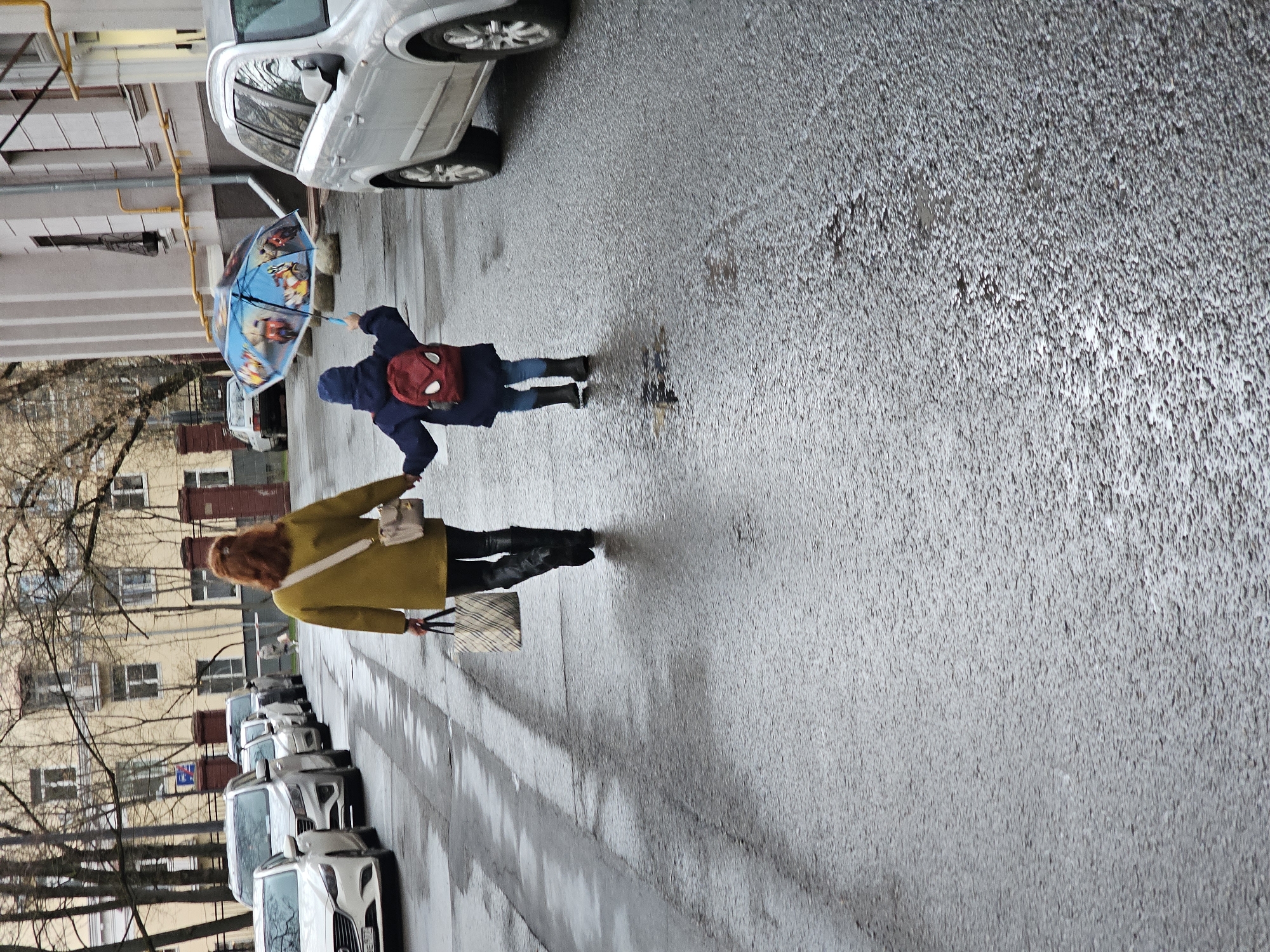 A street scene on a wet, overcast day shows two pedestrians walking on a sidewalk beside a row of parked cars in Khimki, Russia. To the left are older buildings and bare trees. One pedestrian, walking closer to the cars, wears a long yellow coat and carries a light-colored handbag and a shopping bag. The other pedestrian is a child wearing a dark blue jacket, blue jeans, and black boots, carrying a red backpack with white web details, and holding a blue and red children's umbrella. Parked cars line the street on the left of the frame. Behind the pedestrians and cars are taller buildings with windows. The ground is wet, indicating recent or ongoing rain.