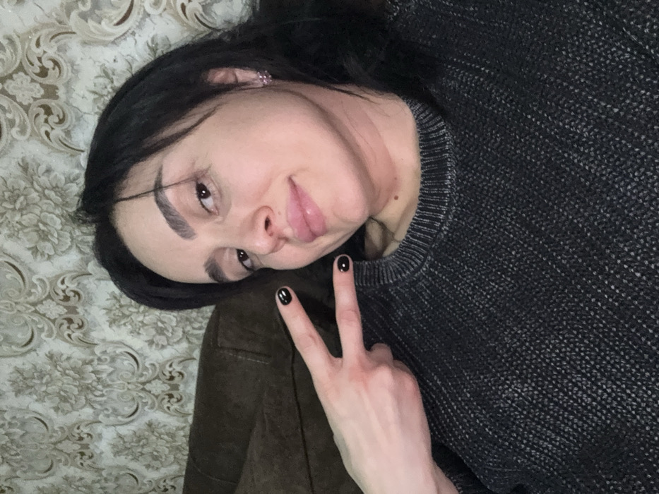 A female subject with dark hair and light complexion is depicted in a close-up, chest-up selfie format. Her left hand, with fingernails painted black, is raised to the left of her head, forming a "V" peace sign with the index and middle fingers extended. She is wearing a dark grey, possibly charcoal, knit sweater with a ribbed collar visible around her neck. Her expression is a slight smile, with full, glossy lips. A small, light-colored stud earring is visible in her right ear. The background consists of a wall covered in patterned wallpaper featuring a light floral or damask design in tones of beige and cream. A section of a dark brown upholstered couch or furniture item is visible behind her left shoulder. The specific location within the city of Bolnisi, Georgia, cannot be determined from the provided image.