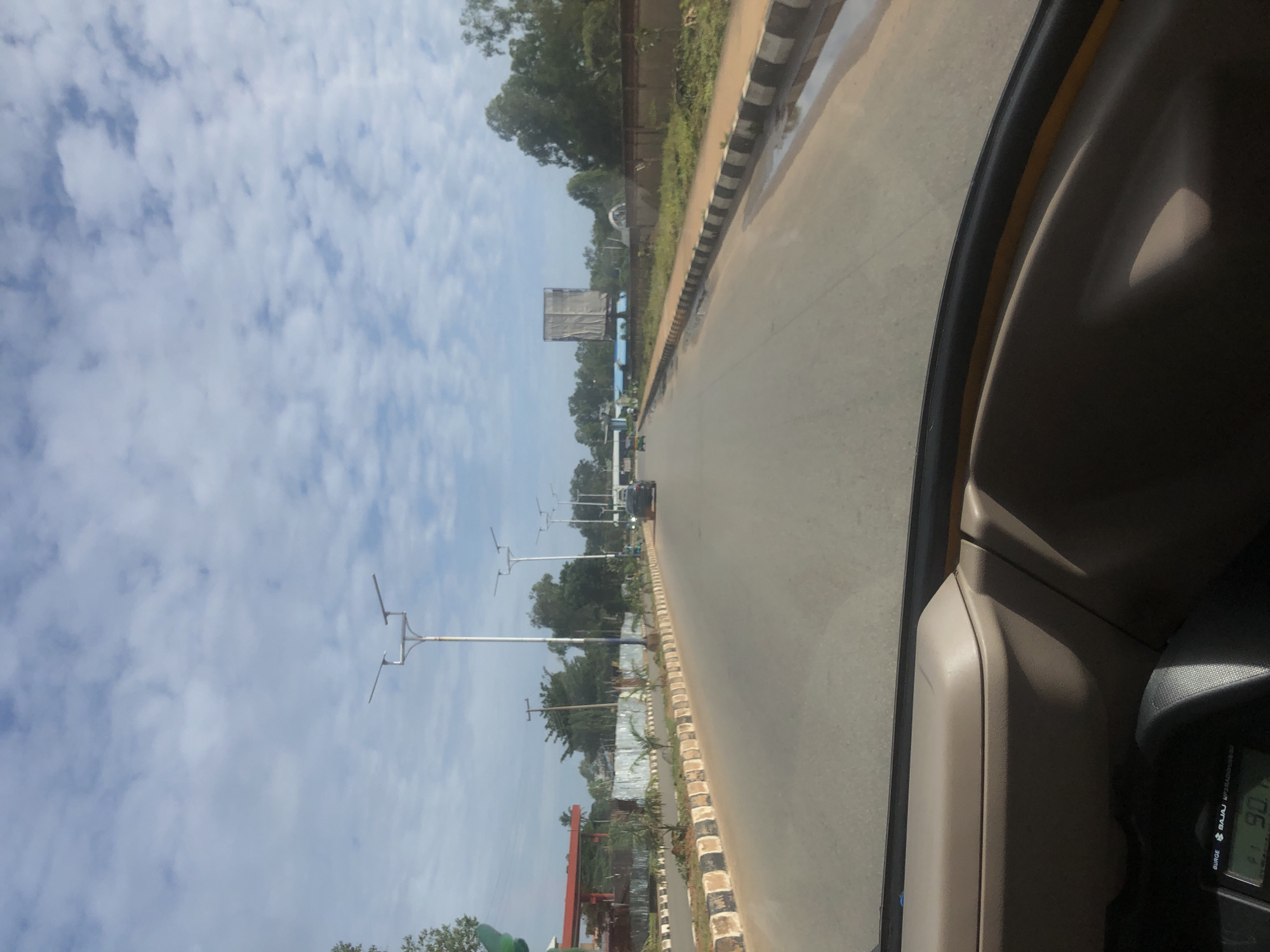 The image depicts a forward-facing view from inside a vehicle traveling on a paved road in Barakin Lerawi, Nigeria. The road is divided by a median strip with black and white striped borders and lined with small palm trees. Streetlights are visible along the roadside. On the left side of the road, there is a gas station with a red canopy and a fence. Further down the road, on the right, a billboard covered with a light-colored tarp is visible. The sky is partly cloudy. Several vehicles, including a dark sedan and a green auto-rickshaw, are present on the road. The interior of the vehicle, including a portion of the dashboard with a digital display showing "90," is visible in the foreground.