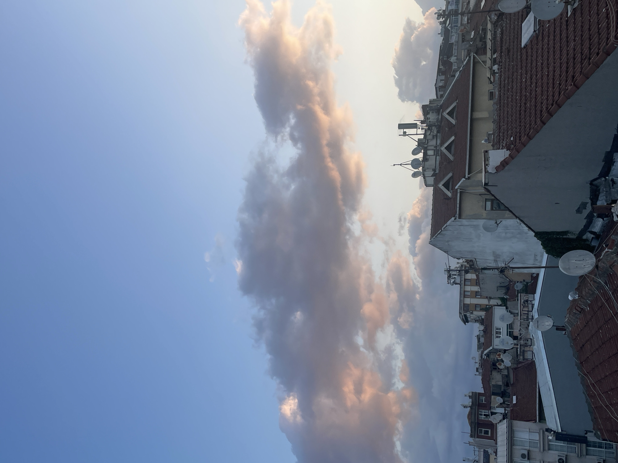 The image captures a rooftop view of Kadıköy, Turkey, with a sky filled with soft, pink-tinged clouds. In the foreground, various rooftops with terracotta tiles are visible, some with satellite dishes. Buildings of different architectural styles are clustered together, some appearing aged and others more modern. The sky is a gradient of light blue transitioning to lighter hues near the horizon. The overall impression is a serene urban landscape bathed in the gentle light of either sunrise or sunset.