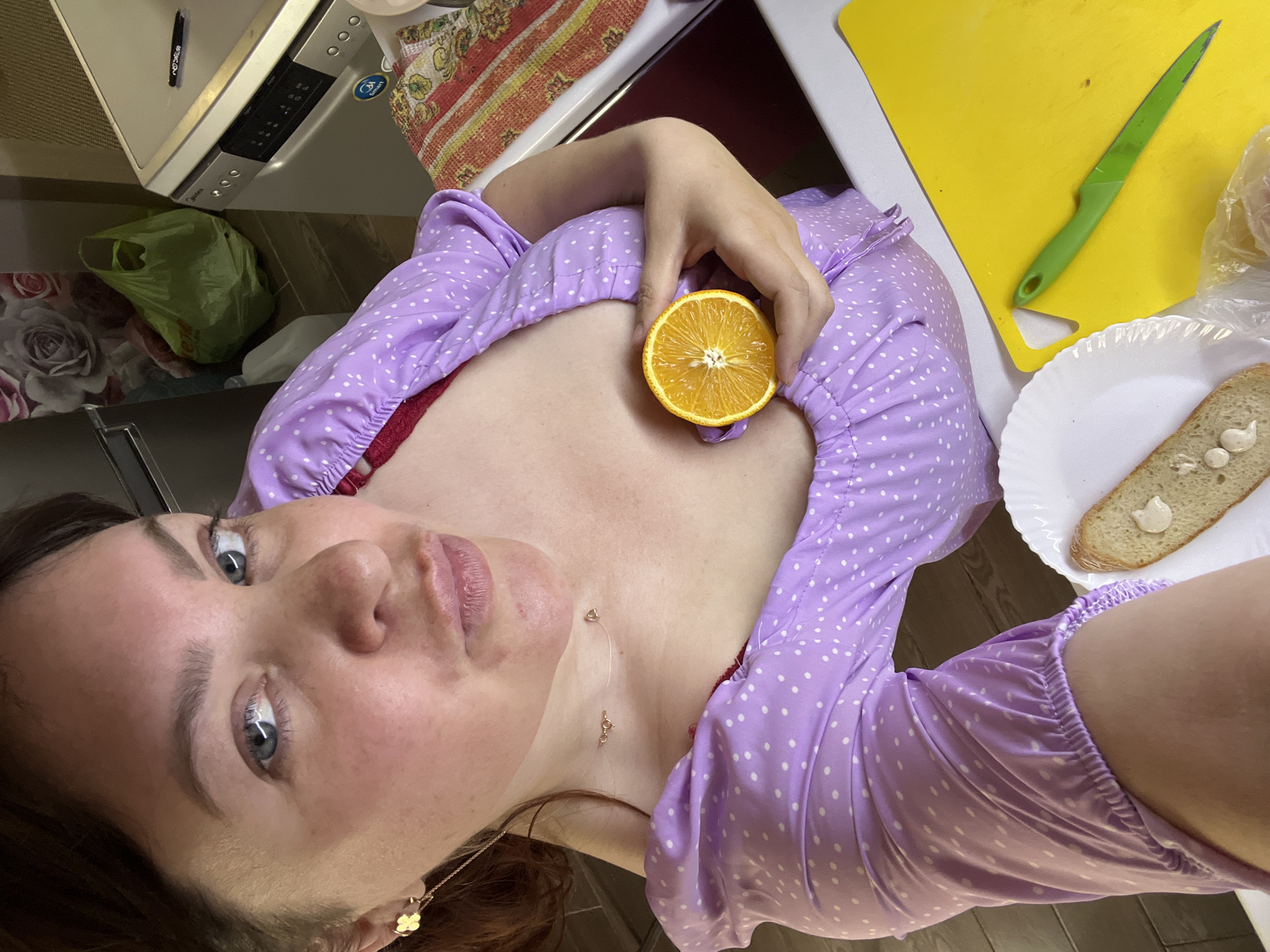 The image shows a woman in a kitchen. She is wearing a light purple polka dot dress with a red undergarment visible. The woman is holding a slice of orange to her chest. In the foreground, there is a white plate with a slice of bread topped with a creamy substance, and a yellow cutting board with a green knife. In the background, a dishwasher is visible, along with a green plastic bag. The lighting suggests it is daytime. The location provided, Noviy, Russia, cannot be verified from the visual information in the image.