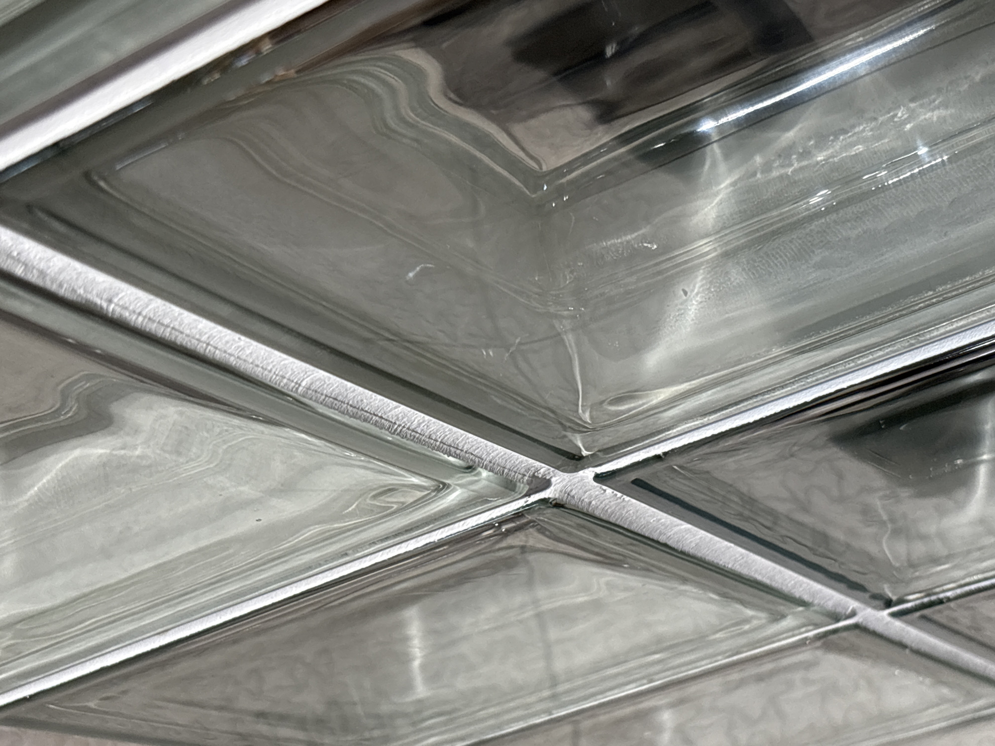 The image is a close-up, abstract view of glass blocks, likely part of a wall or partition. The glass has a textured, rippled surface that distorts and reflects light, creating patterns of light and shadow. The overall color palette is muted grays and whites, with hints of the underlying material within the glass. There are no people or discernible objects within the frame, making the image entirely focused on the texture and light play of the glass blocks. The specific location in Saint Petersburg, Russia, is not identifiable from the visual information provided in the image.