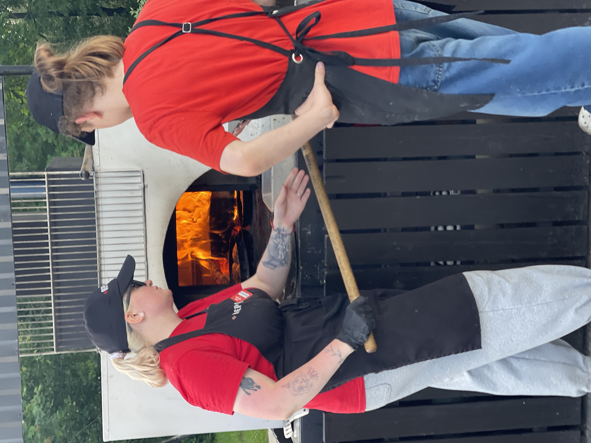 Two individuals, one wearing a black baseball cap, sunglasses, and a red t-shirt with a black apron, and the other wearing a black baseball cap and a red t-shirt with a black apron, are standing in front of a wood-fired pizza oven. The person on the left, with blonde hair in a ponytail, is holding a long wooden-handled tool and a white rectangular pan. The person on the right, with long brown hair tied back, is facing away from the camera and has their hand on their hip. Flames are visible inside the oven. The environment appears to be outdoors, with trees and foliage in the background. The subjects are positioned in what looks like a food service area.
