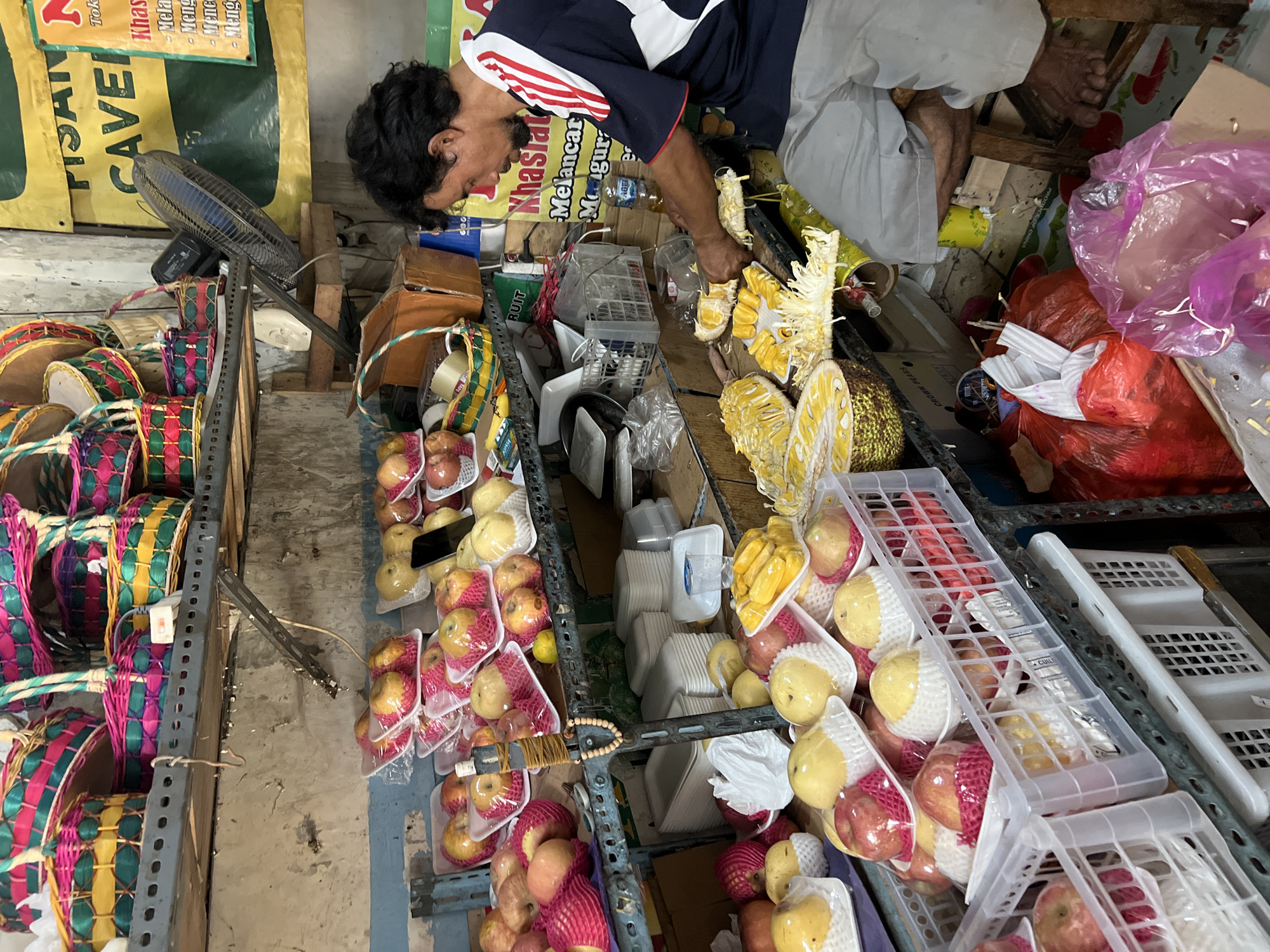 In Gunung Putri, Indonesia, a man is seen preparing jackfruit. He is sitting on a wooden stool, wearing a blue and white striped shirt and light gray shorts. His hands are engaged in cutting and separating the yellow fruit segments, which are arranged on a white plate in front of him. Several large pieces of jackfruit, both cut and whole, are visible on a table. The scene appears to be a market stall, with shelves stocked with various fruits, including apples and pears, neatly packaged in plastic. Colorful woven baskets are also displayed on a higher shelf. In the background, a fan is positioned on a shelf, and a yellow banner with Indonesian text is partially visible. The environment is cluttered, with plastic bags, containers, and other items scattered around. The lighting suggests an indoor setting, possibly under artificial light, with natural light contributing from an unseen source.