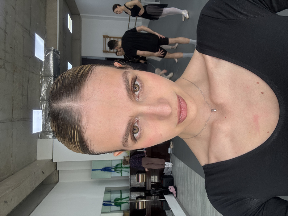 The image depicts the interior of a dance studio, location Unknown, Unknown. In the foreground, a female with light brown hair pulled back from a central part, wearing a black long-sleeved leotard and a silver pendant necklace, looks directly at the camera. A faint reddish mark is visible on her upper left chest.
The background reveals a spacious room with white walls, a dark grey floor, and an exposed concrete ceiling featuring rectangular LED light panels and ventilation ducts. On the left side, a large mirror reflects a dark wooden cabinet or piano topped with books. A shorter individual, wearing a dark patterned top, light brown pants, and white sneakers, is partially visible near this furniture. A pair of pink ballet pointe shoes rests on the floor nearby.
On the right side of the background, four additional individuals are present. One female, dressed in a black leotard, short black skirt, white tights, and pointe shoes, stands with her hands on her hips. Adjacent to her, a male wears a black t-shirt, black shorts, and grey socks. Two other individuals are partially visible further back. A wooden ballet barre is mounted along the white wall. The individuals in the background appear to be in a relaxed or paused state, not actively dancing or interacting with the foreground subject.