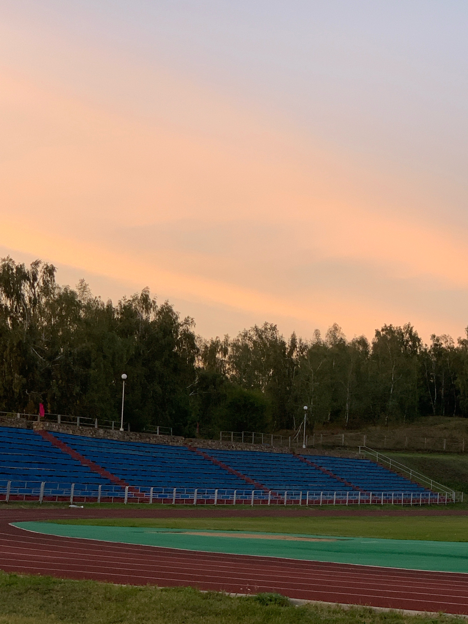 The image captures an outdoor stadium at dusk. The sky displays a gradient of pale orange and soft purple hues, with wispy clouds. A dense line of trees with dark green foliage forms a backdrop behind the stadium seating. The stadium itself features rows of blue seats with red accents, bordered by metal railings. In the foreground, a running track with red and green lanes is visible, bordered by grass. A single lamppost stands near the treeline, and a small pink flag is planted on top of the stadium's stone wall. The scene is serene and devoid of people, suggesting the stadium is empty. The image does not contain any specific landmarks that would definitively place it within the city of Kapyĺ, Belarus.