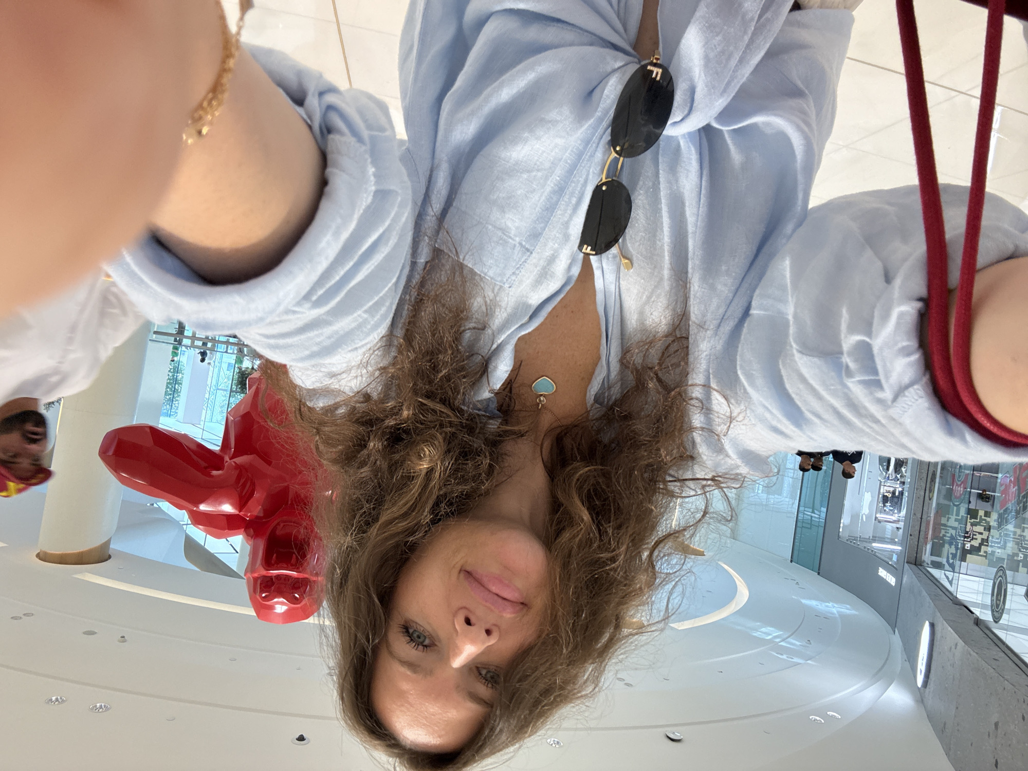 The image shows a woman taking a selfie in a modern, well-lit indoor shopping mall. She is wearing a light blue, oversized button-down shirt with her hair down and curly. Around her neck, she has a necklace with a turquoise heart-shaped pendant, and a pair of sunglasses is hooked onto her shirt. She is holding a red strap in her left hand. In the background, there is a large, bright red, abstract sculpture of a gorilla. To the right of the sculpture, a man wearing a white shirt and a red cap is visible. In the further background, there are shops with glass facades and other mall-goers. The overall environment suggests a high-end retail setting. Based on the distinctive architecture and the presence of the red gorilla sculpture, this scene is likely at The Dubai Mall in Dubai, United Arab Emirates.