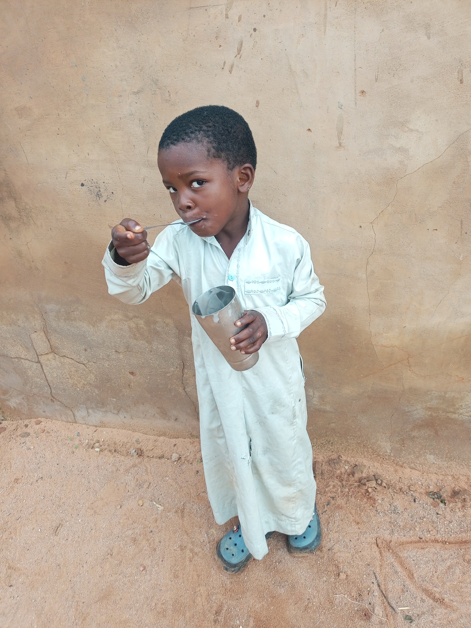 A young Black boy stands against a rough, tan, stucco wall. He is wearing a light blue, long-sleeved robe with embroidered trim around the collar and down the front opening. The robe is slightly dirty at the hem. He is wearing blue Crocs. The boy is holding a metal cup in his left hand and a spoon in his right hand, which he is using to eat from the cup. His gaze is directed at the viewer. The ground is sandy and reddish-brown, with scattered small pebbles and debris. This scene appears to be outdoors in a rural or undeveloped area, consistent with what might be found in Tempure, Nigeria.